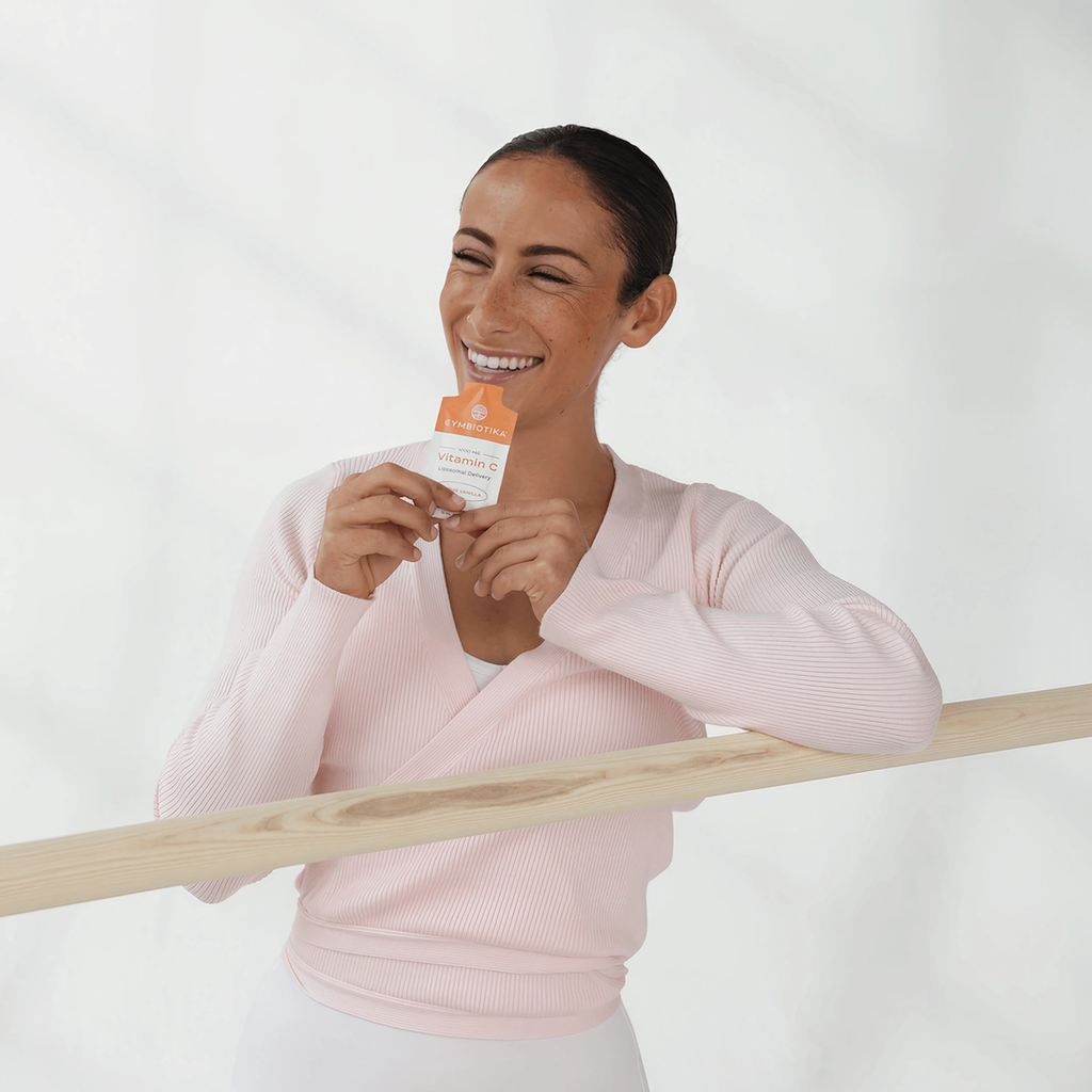 Smiling woman in a pale pink top holds a Cymbiotika Liposomal Vitamin C packet beside a wooden barre.