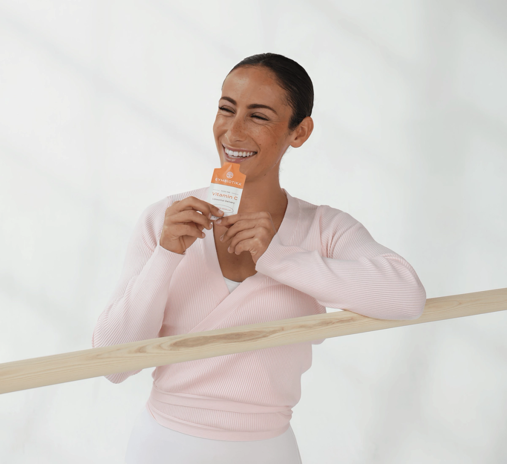 Smiling woman in a pale pink top holds a Cymbiotika Liposomal Vitamin C packet beside a wooden barre.