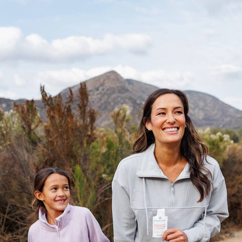 Smiling woman and young girl hold hands on a mountain trail; the woman holds a small supplement packet.