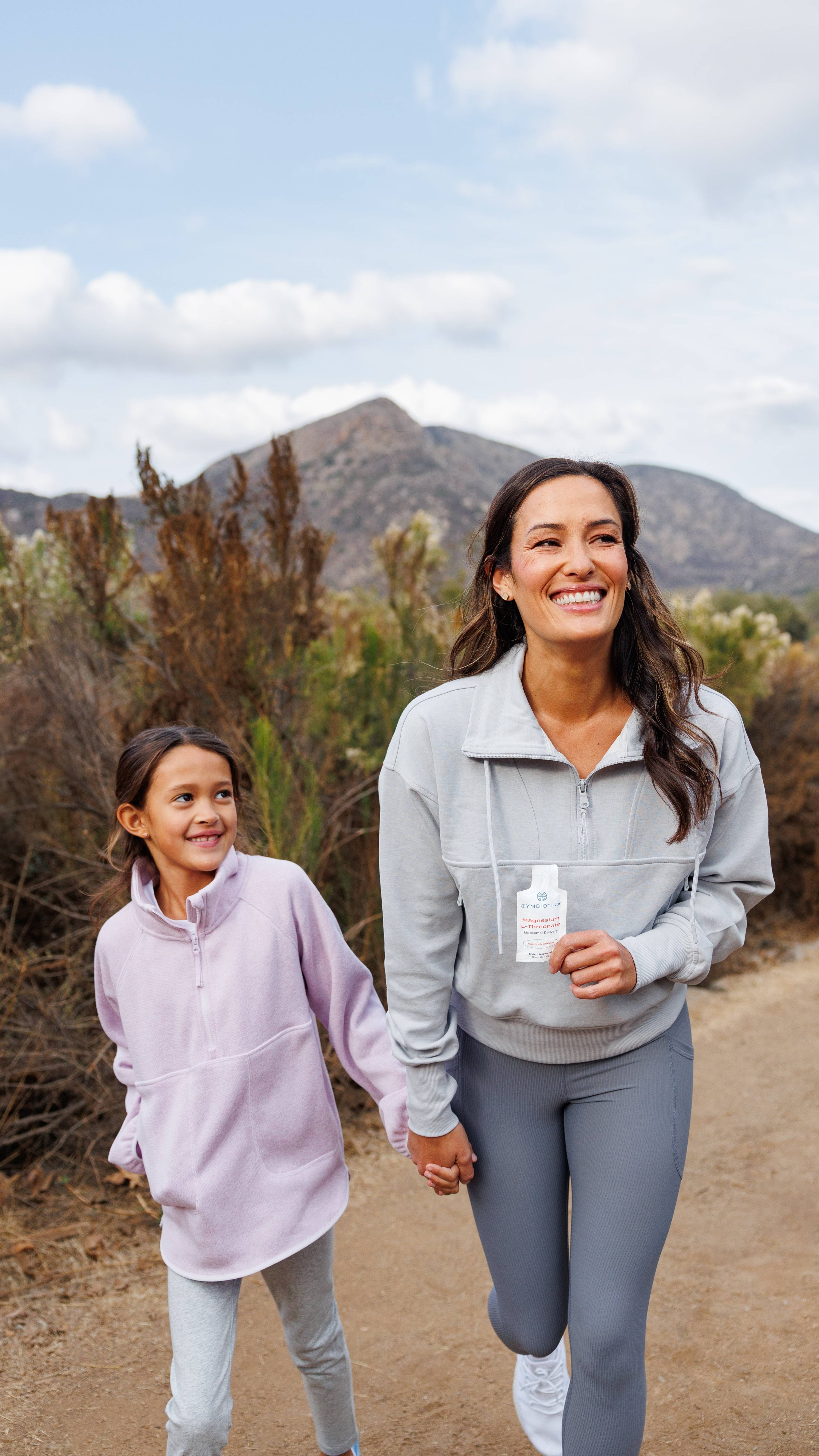 Smiling woman and young girl hold hands on a mountain trail; the woman holds a small supplement packet.