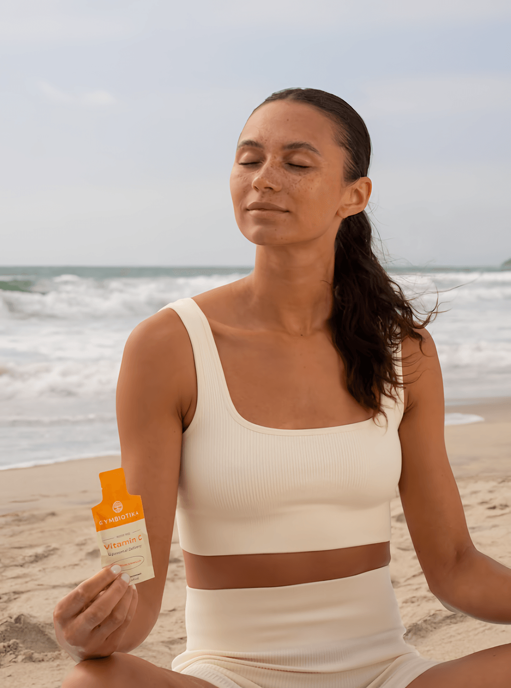 A woman sits on a beach with her eyes closed, holding an orange Vitamin C packet.