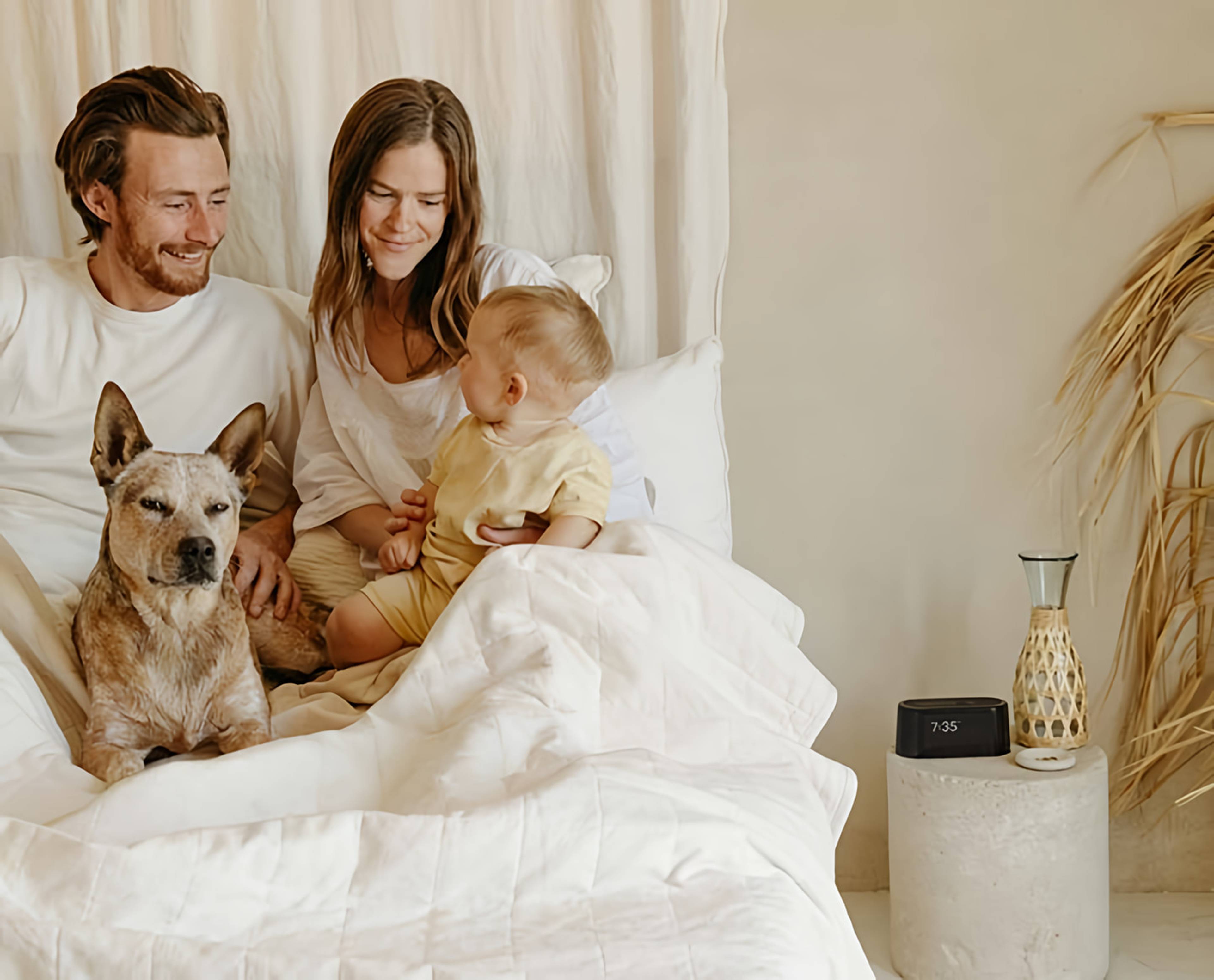 Couple, baby, and dog sitting in a white bed beside a digital alarm clock.
