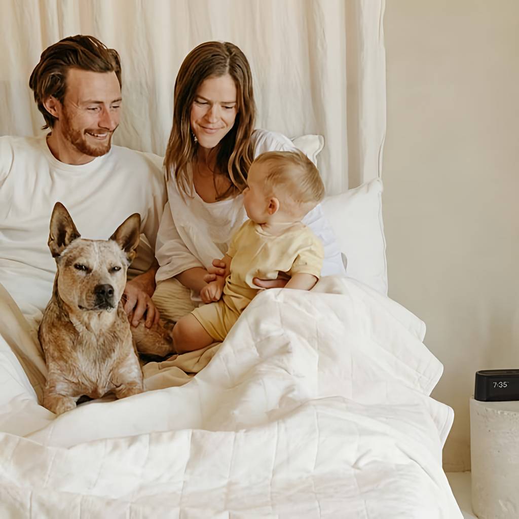 Couple, baby, and dog sitting in a white bed beside a digital alarm clock.