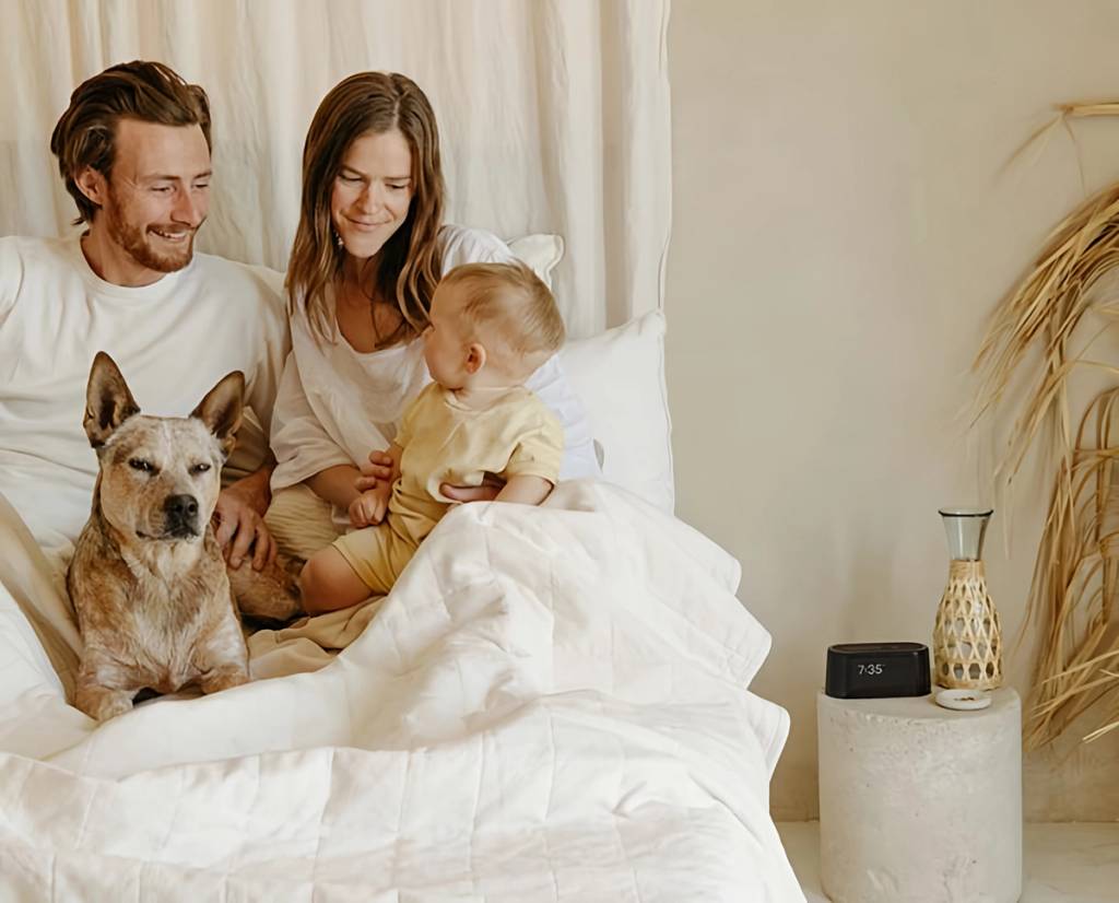 Couple, baby, and dog sitting in a white bed beside a digital alarm clock.
