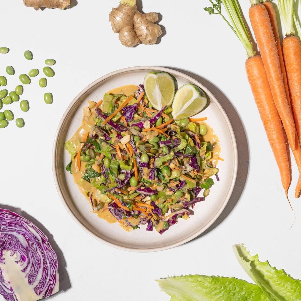 A bowl of colorful chopped salad with lime wedges, surrounded by carrots, ginger, edamame, cabbage, and lettuce.