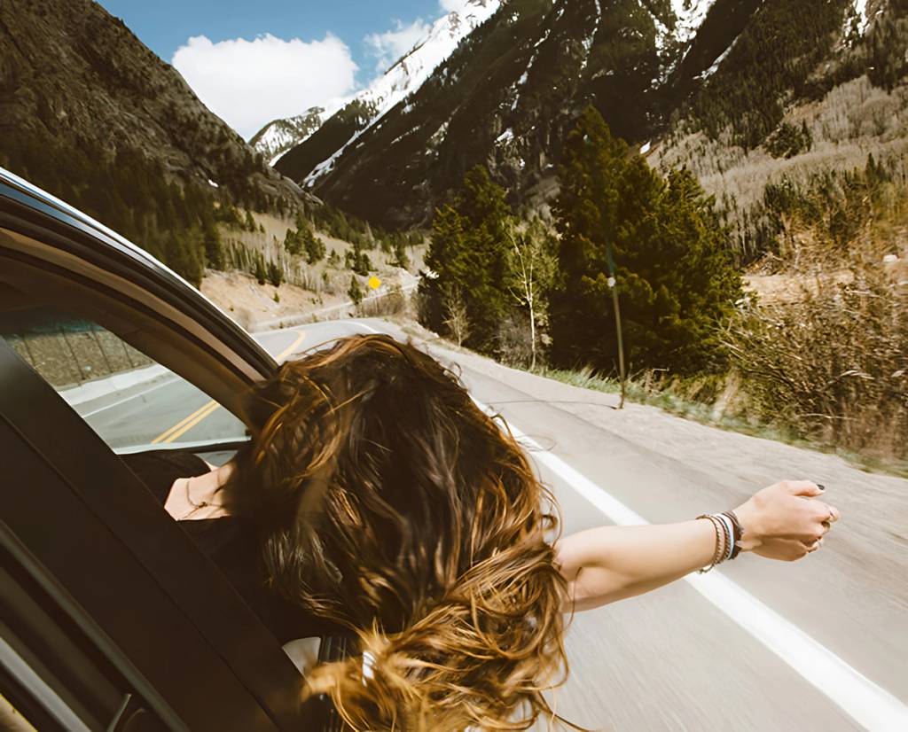 Person with long hair leans out of a car window on a mountain road.