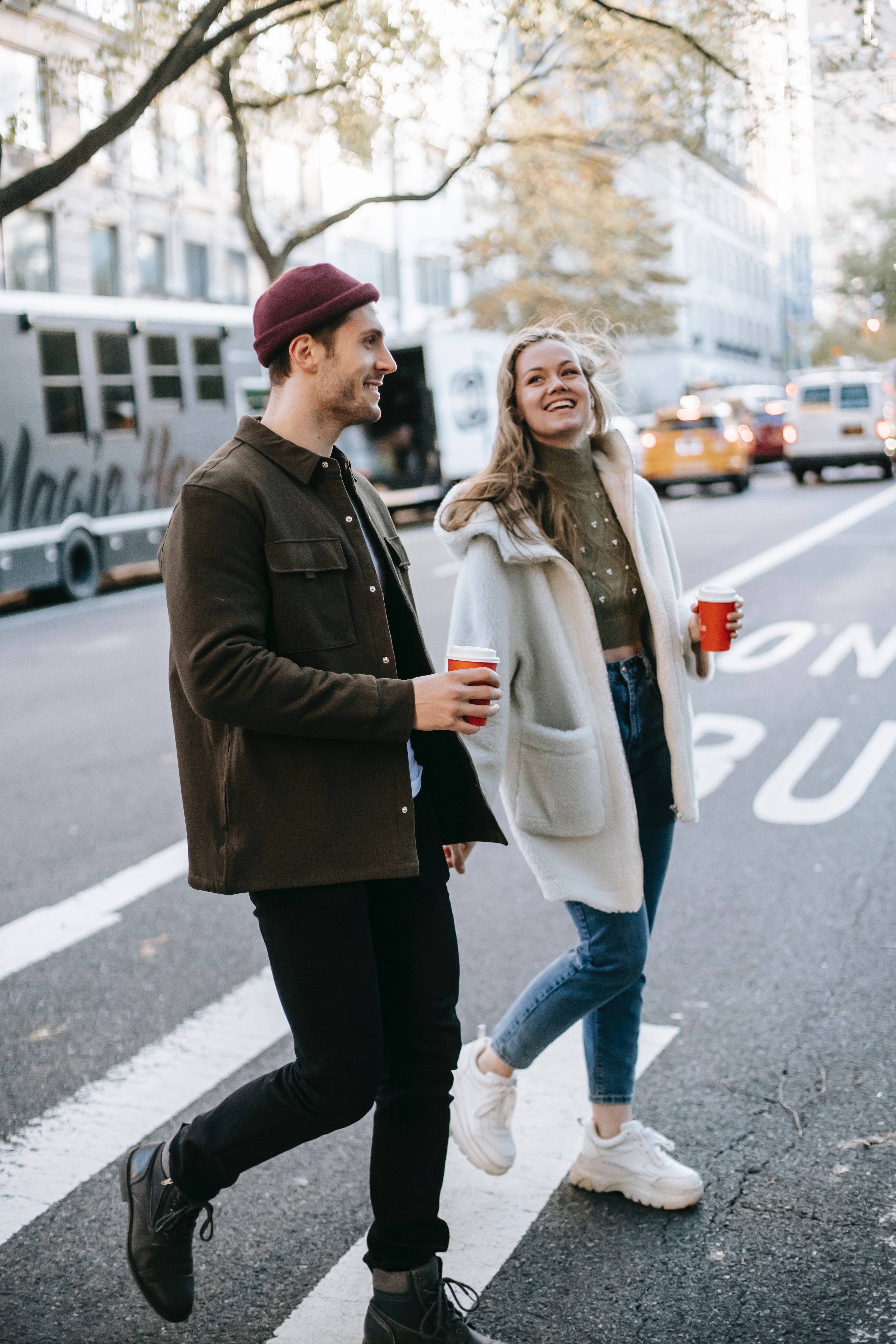 A smiling man and woman walk across a city street, each holding a red cup with a white lid.
