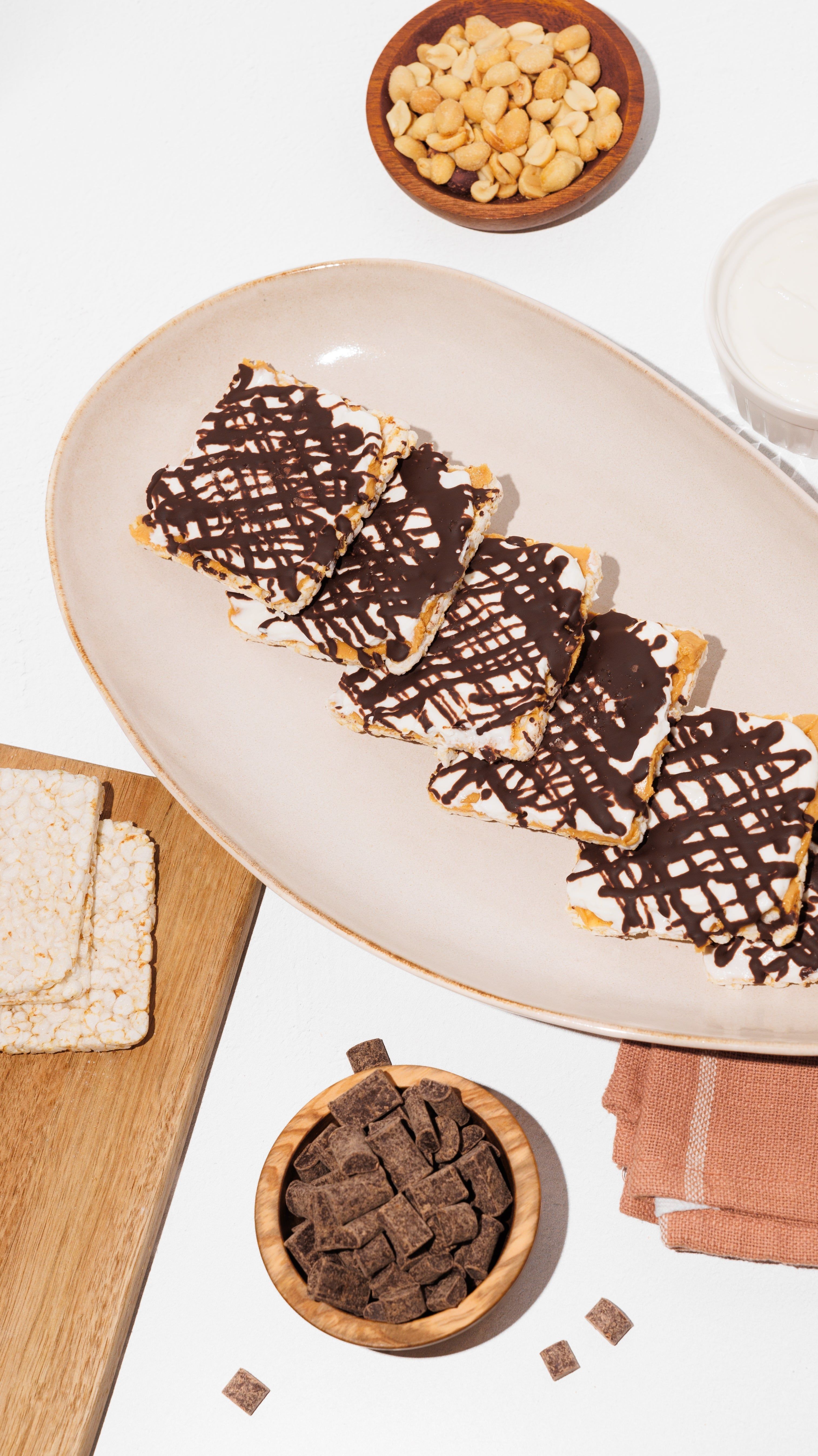 Chocolate-drizzled rice cakes on a plate, with plain rice cakes, peanuts, and chocolate pieces nearby.
