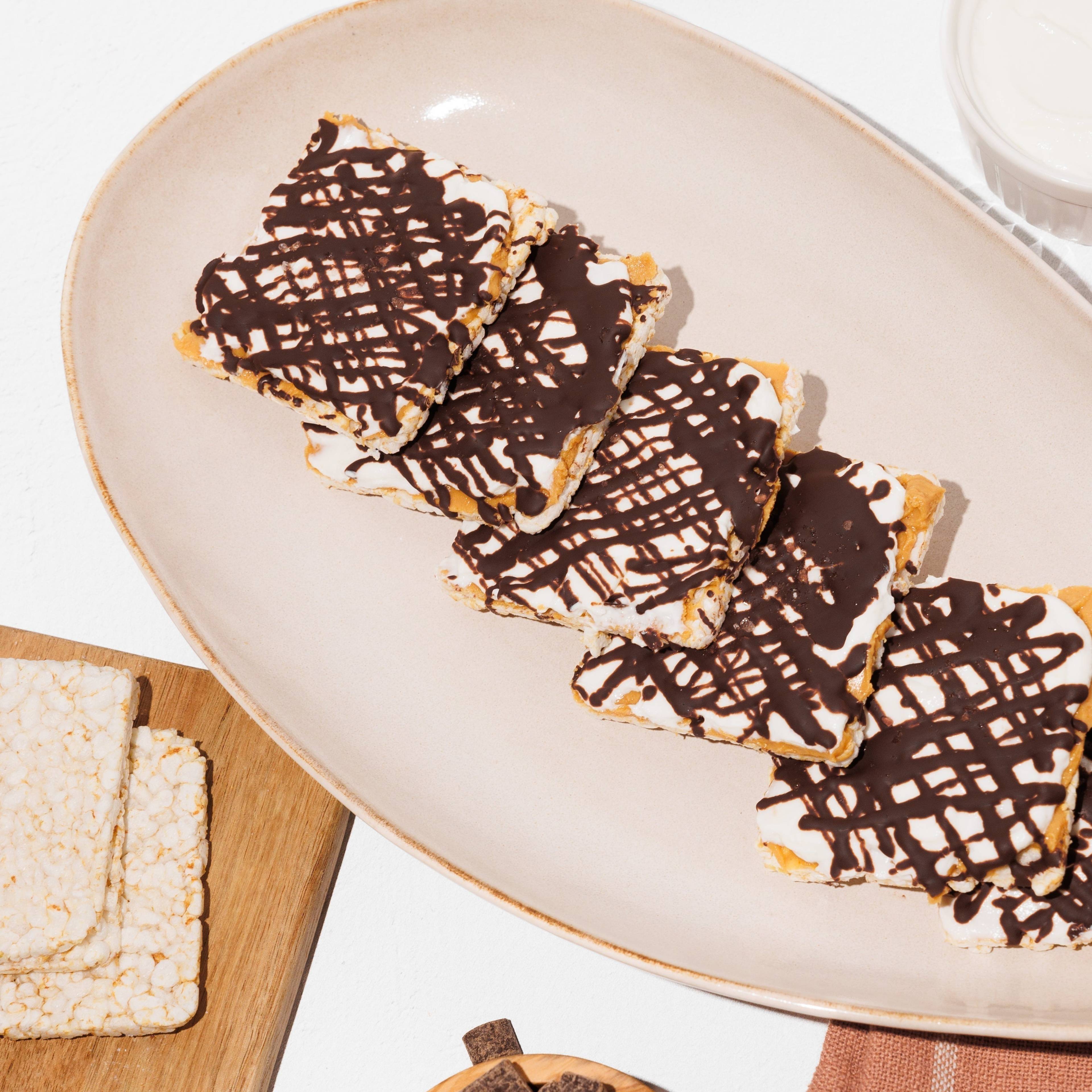 Chocolate-drizzled rice cakes on a plate, with plain rice cakes, peanuts, and chocolate pieces nearby.