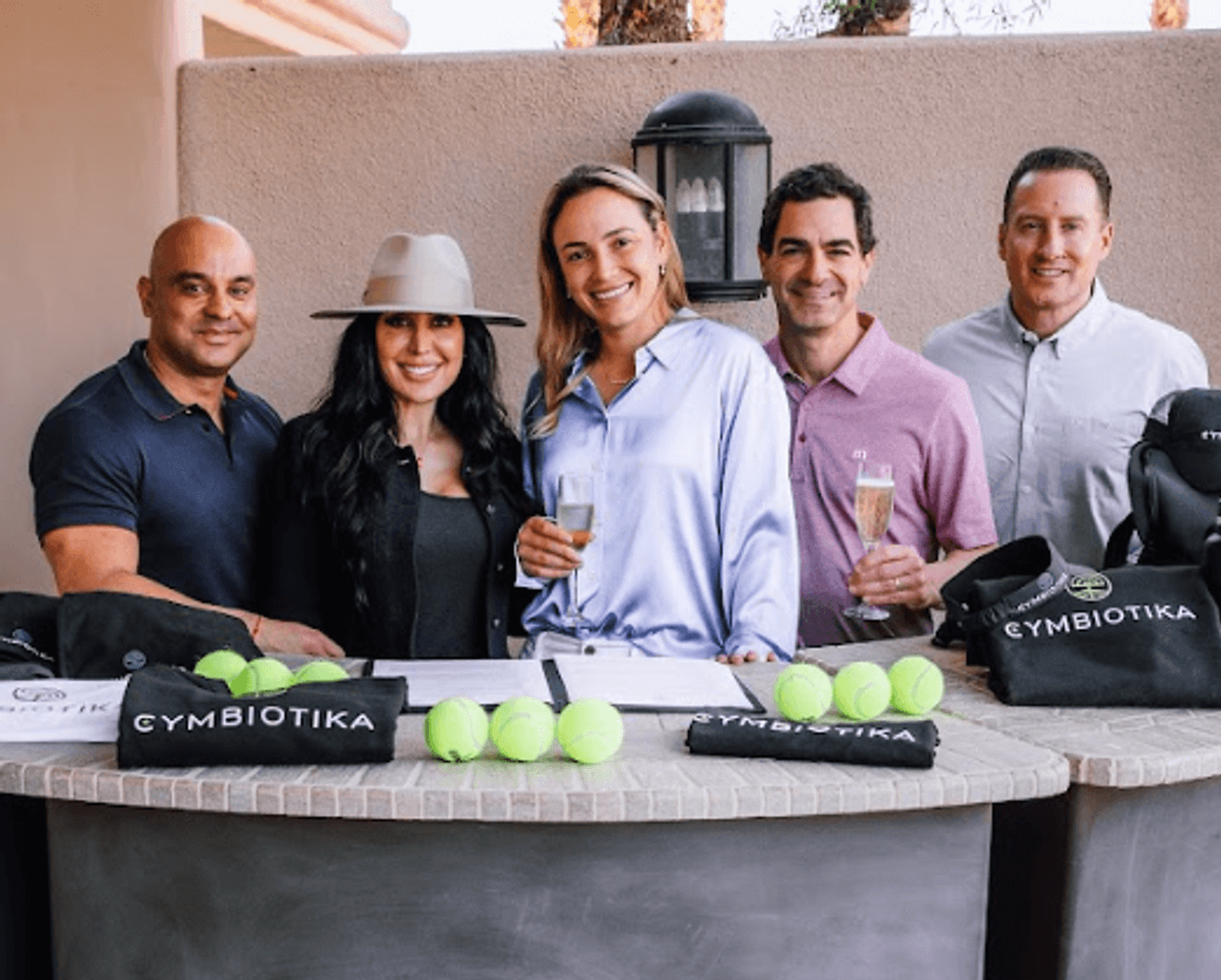 Five adults stand behind a table with tennis balls, champagne glasses, and Cymbiotika-branded bags.