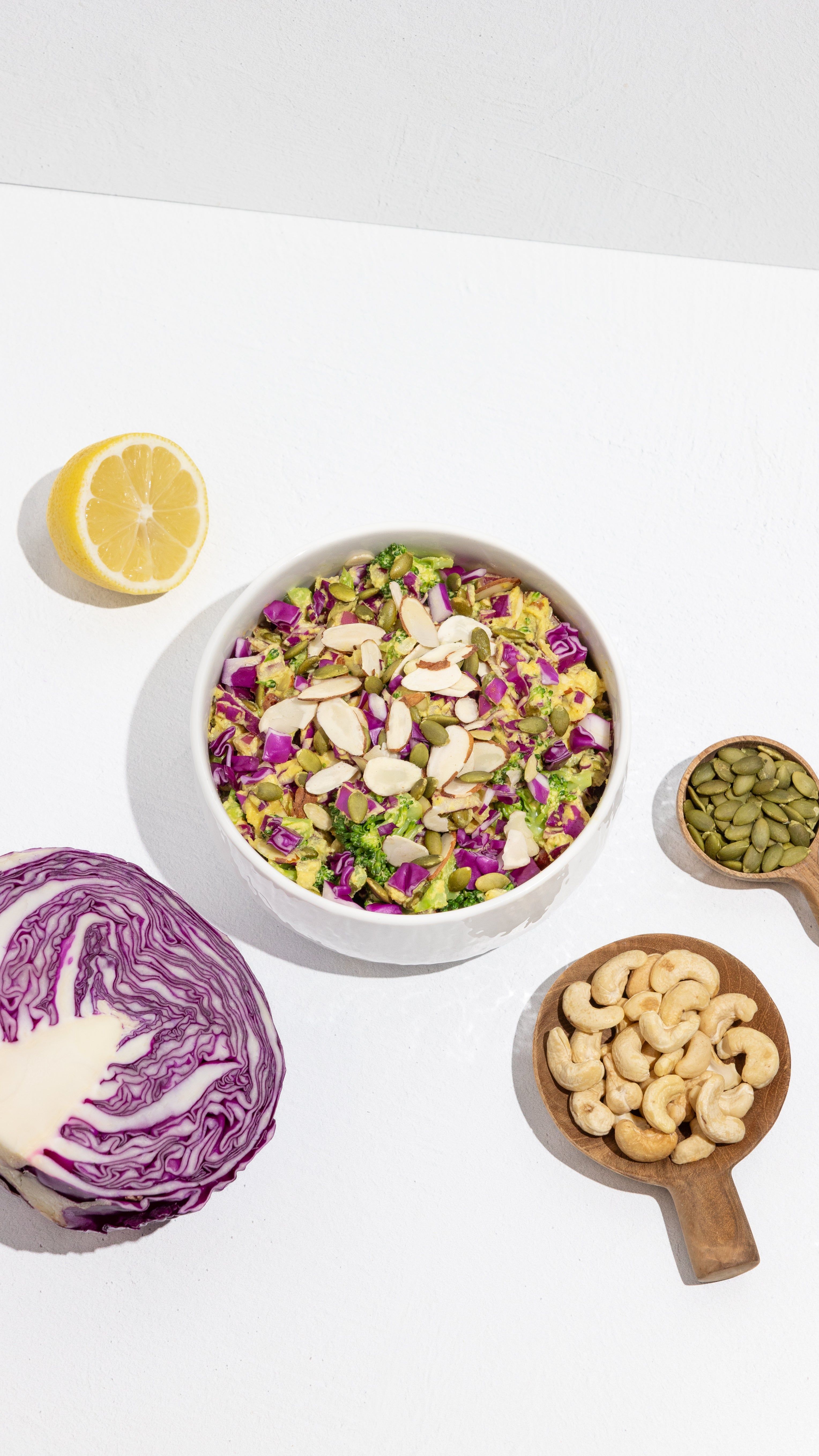 Bowl of broccoli and purple cabbage salad topped with sliced almonds and pumpkin seeds, with lemon, cabbage, and cashews.