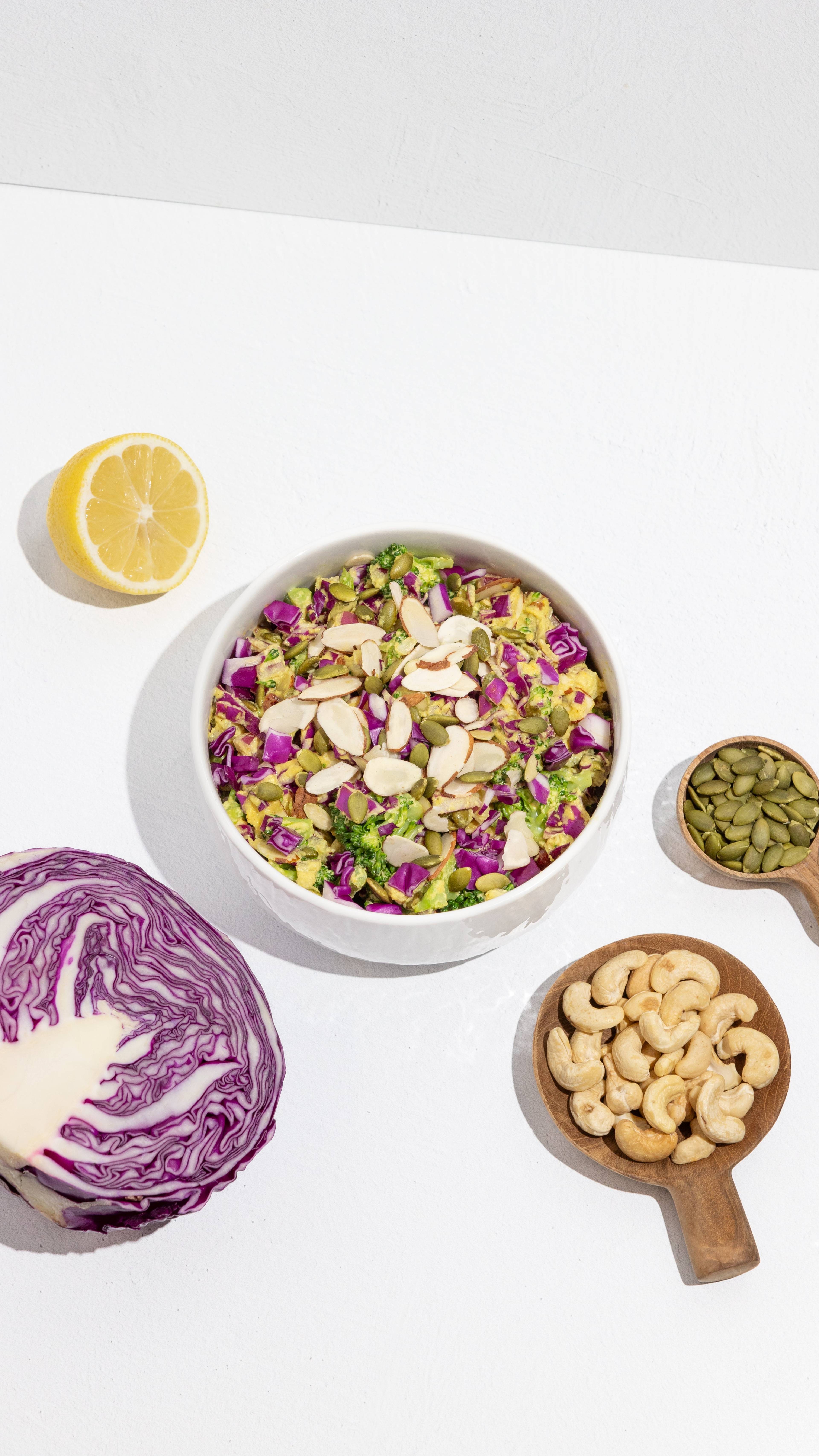 Bowl of broccoli and purple cabbage salad topped with sliced almonds and pumpkin seeds, with lemon, cabbage, and cashews.