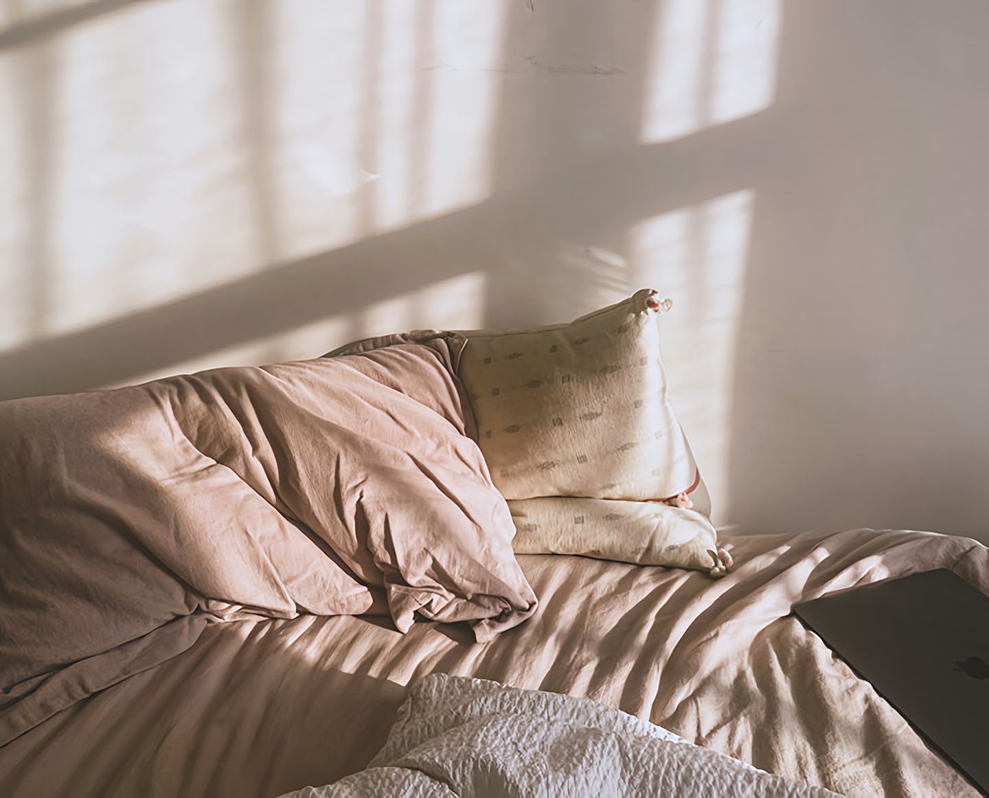 Unmade bed with rumpled pillows, a white blanket, and a closed laptop in window light.