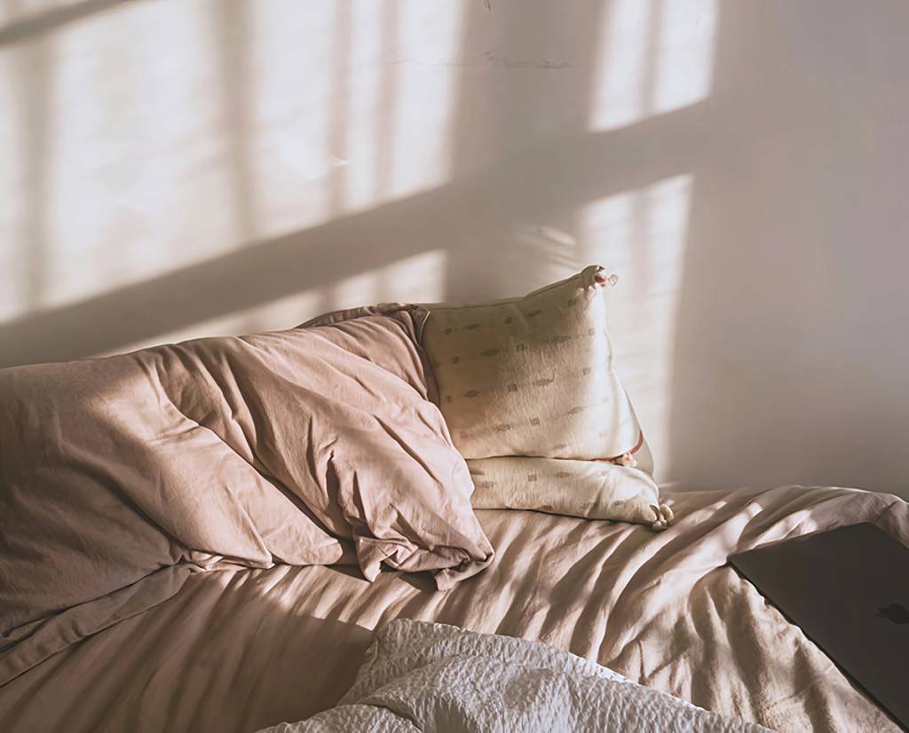 Unmade bed with rumpled pillows, a white blanket, and a closed laptop in window light.