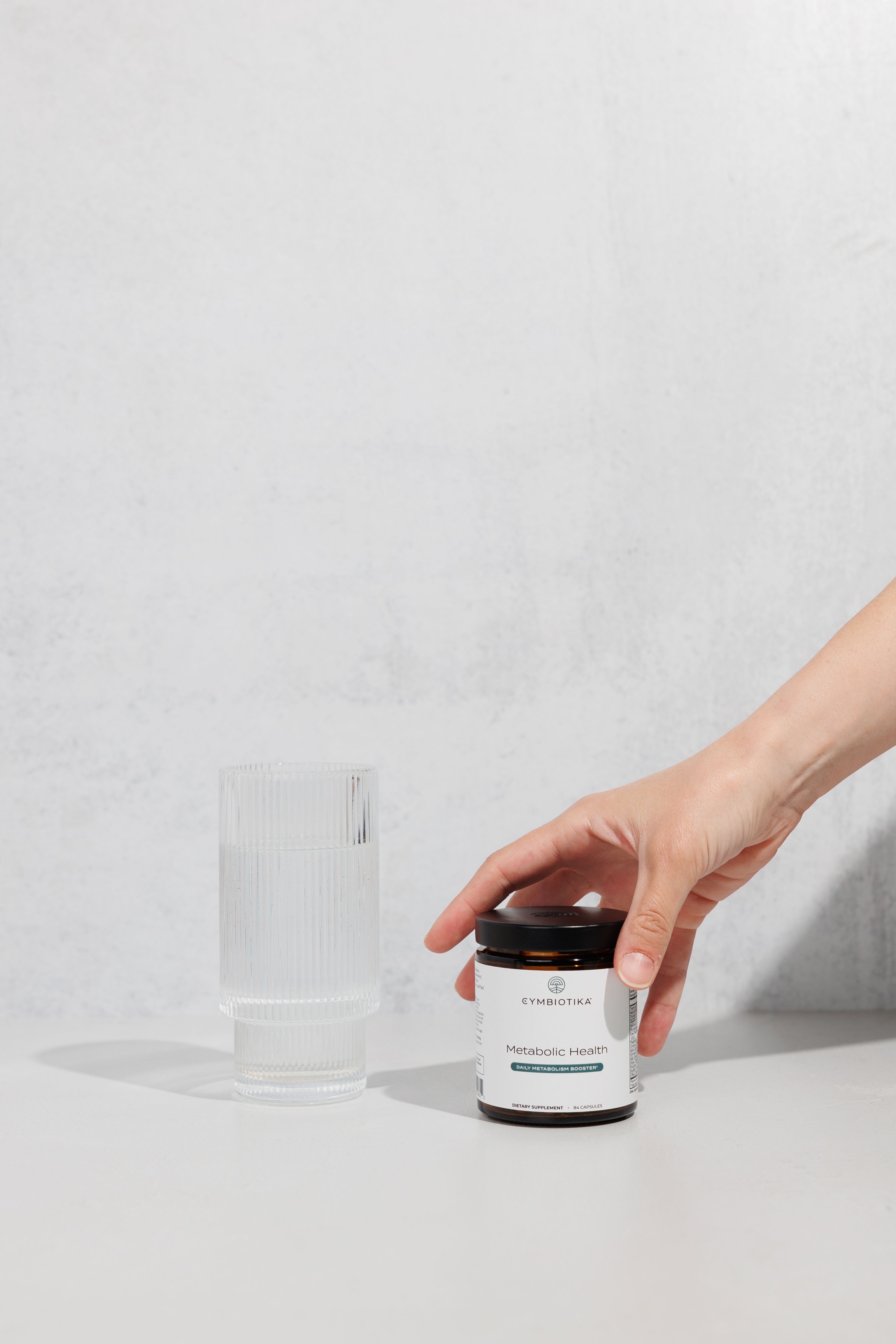 Hand placing a jar of Cymbiotika Metabolic Health beside a ribbed clear glass.