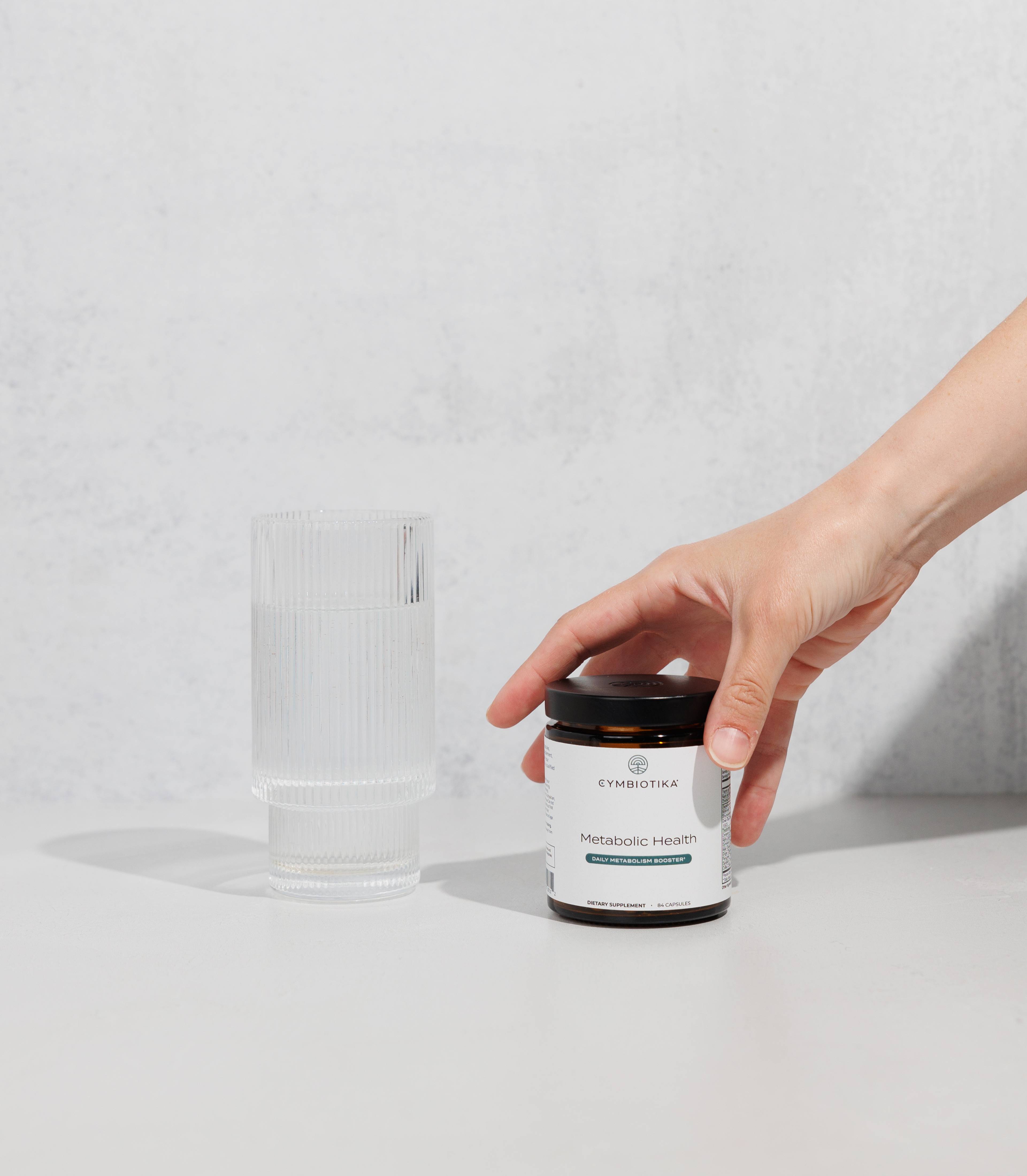 Hand placing a jar of Cymbiotika Metabolic Health beside a ribbed clear glass.