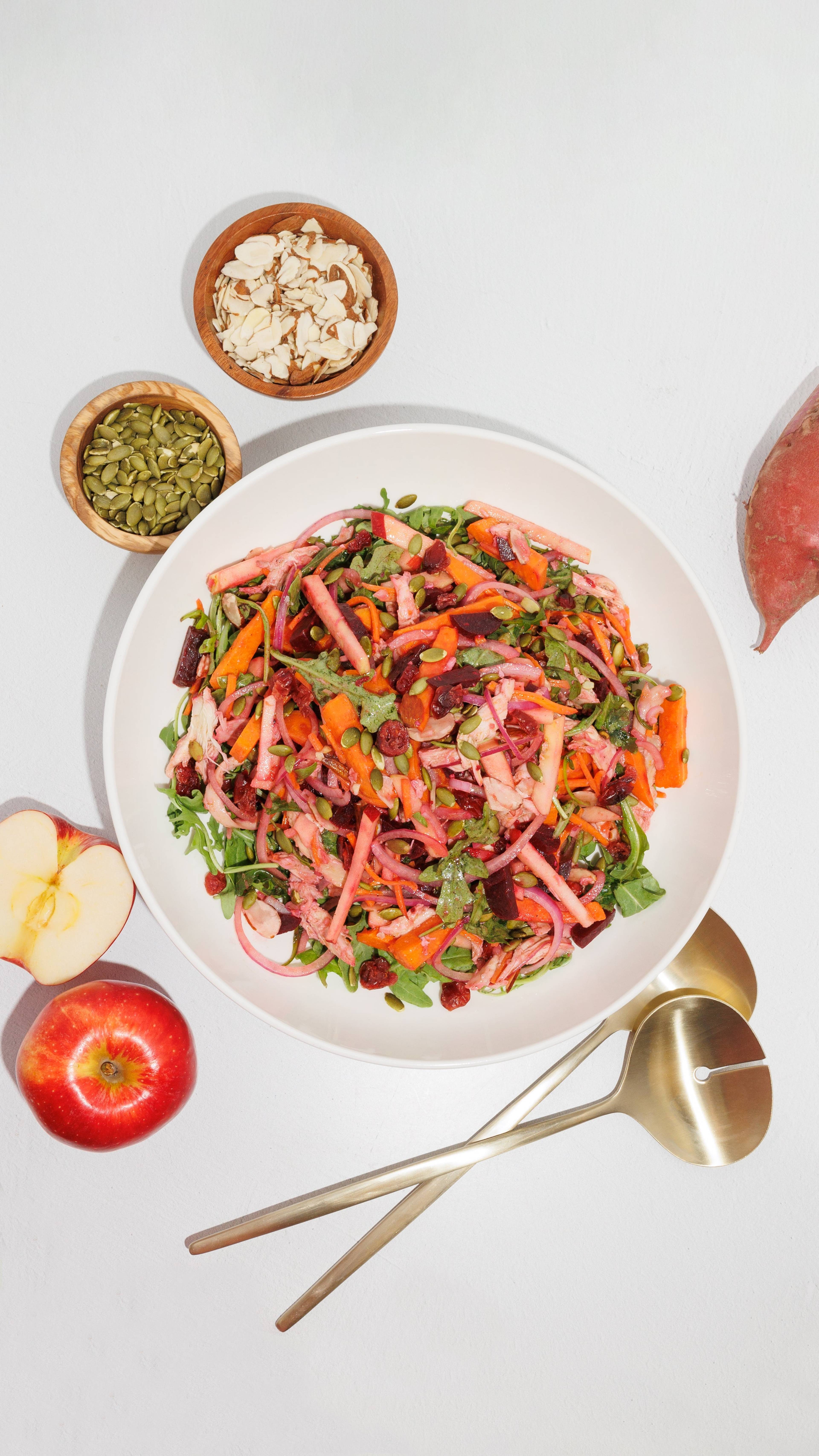 A bowl of chopped salad with leafy greens, colorful vegetable strips, and seeds, with dishes of seeds and sliced almonds nearby.