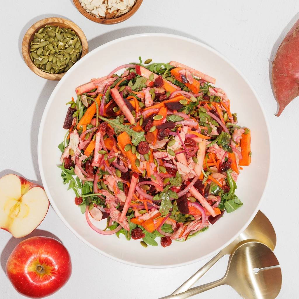 A bowl of chopped salad with leafy greens, colorful vegetable strips, and seeds, with dishes of seeds and sliced almonds nearby.