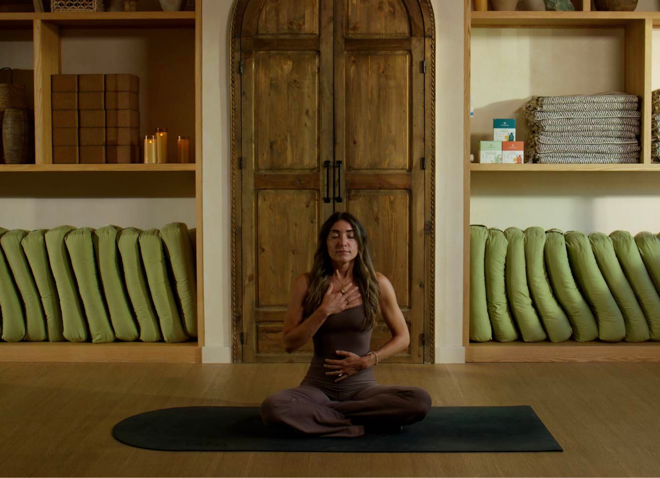 Woman sitting cross-legged with her eyes closed and hands on her chest and abdomen on a dark mat in a wood-paneled room.