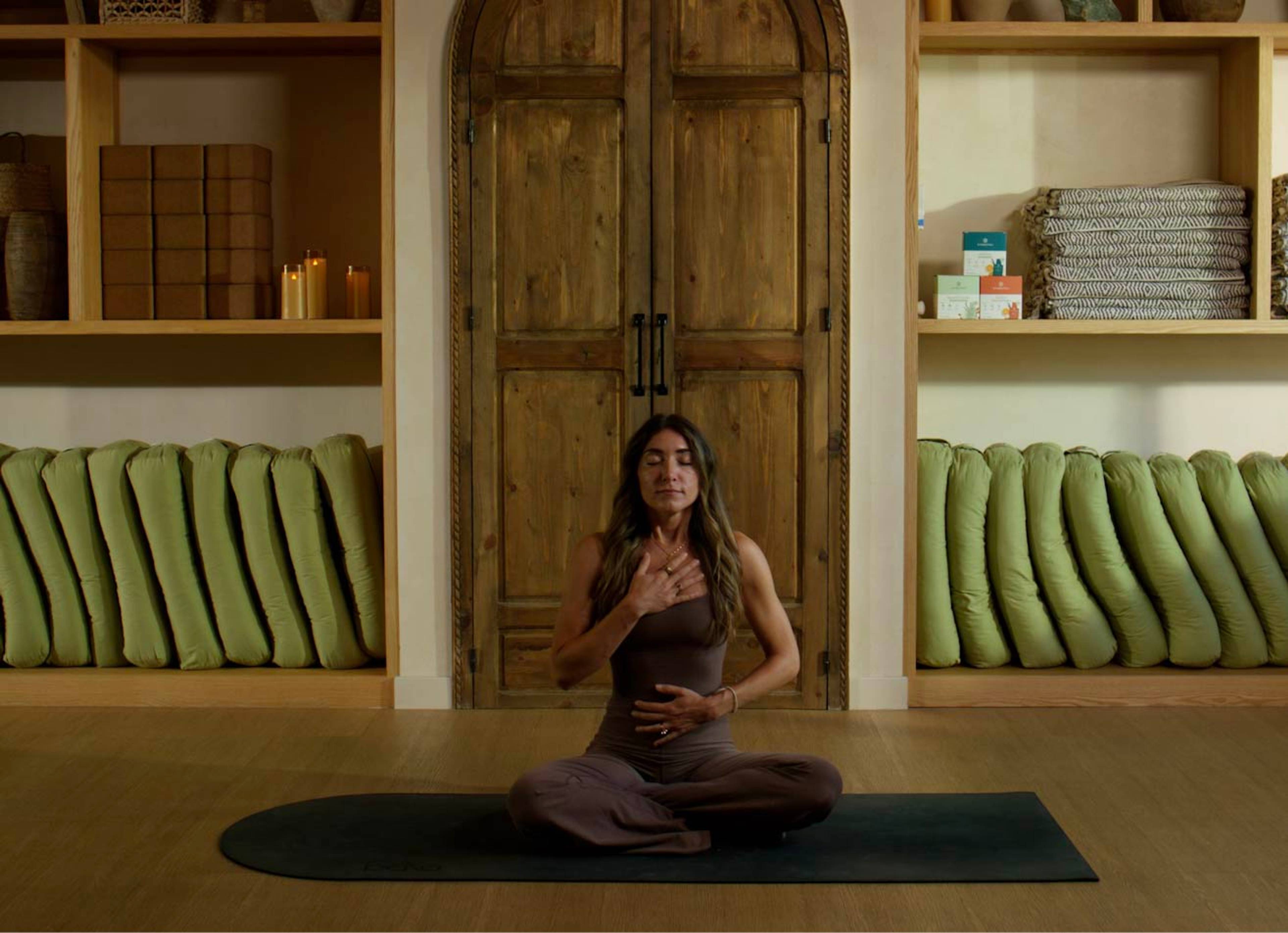 Woman sitting cross-legged with her eyes closed and hands on her chest and abdomen on a dark mat in a wood-paneled room.
