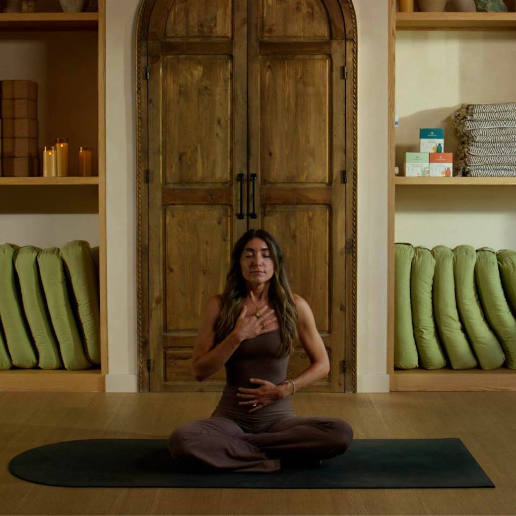 Woman sitting cross-legged with her eyes closed and hands on her chest and abdomen on a dark mat in a wood-paneled room.