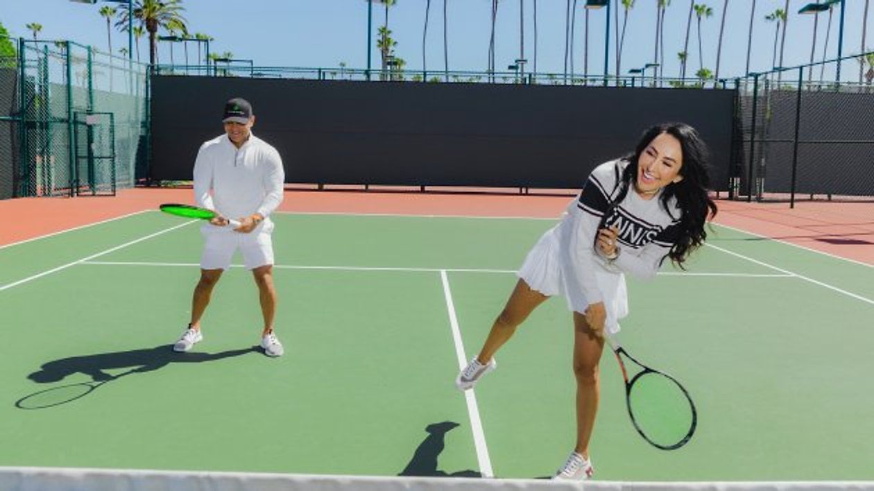 Man and woman playing tennis on an outdoor court lined with palm trees.