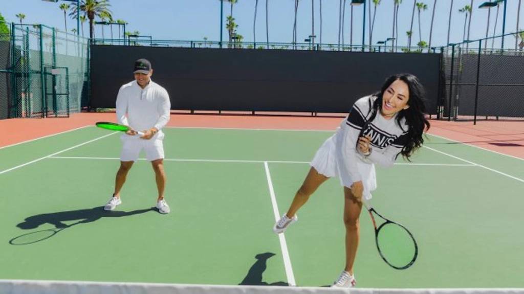 Man and woman playing tennis on an outdoor court lined with palm trees.