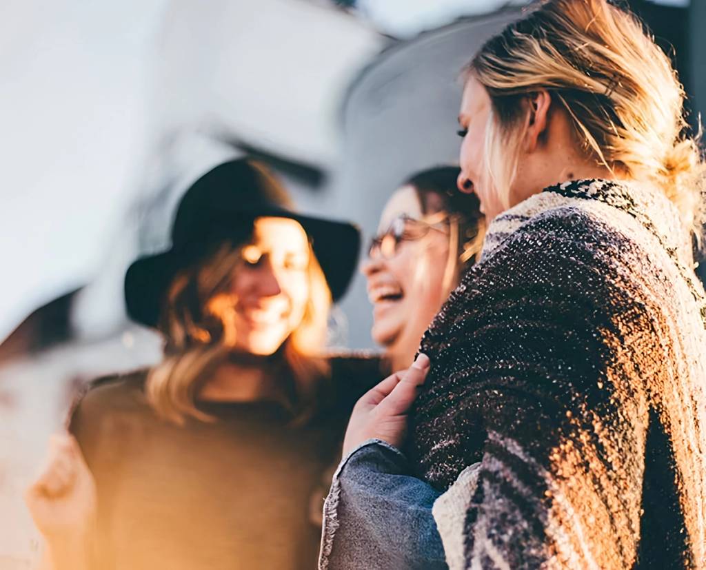 Three women laughing together in bright sunlight; one wears a dark hat.