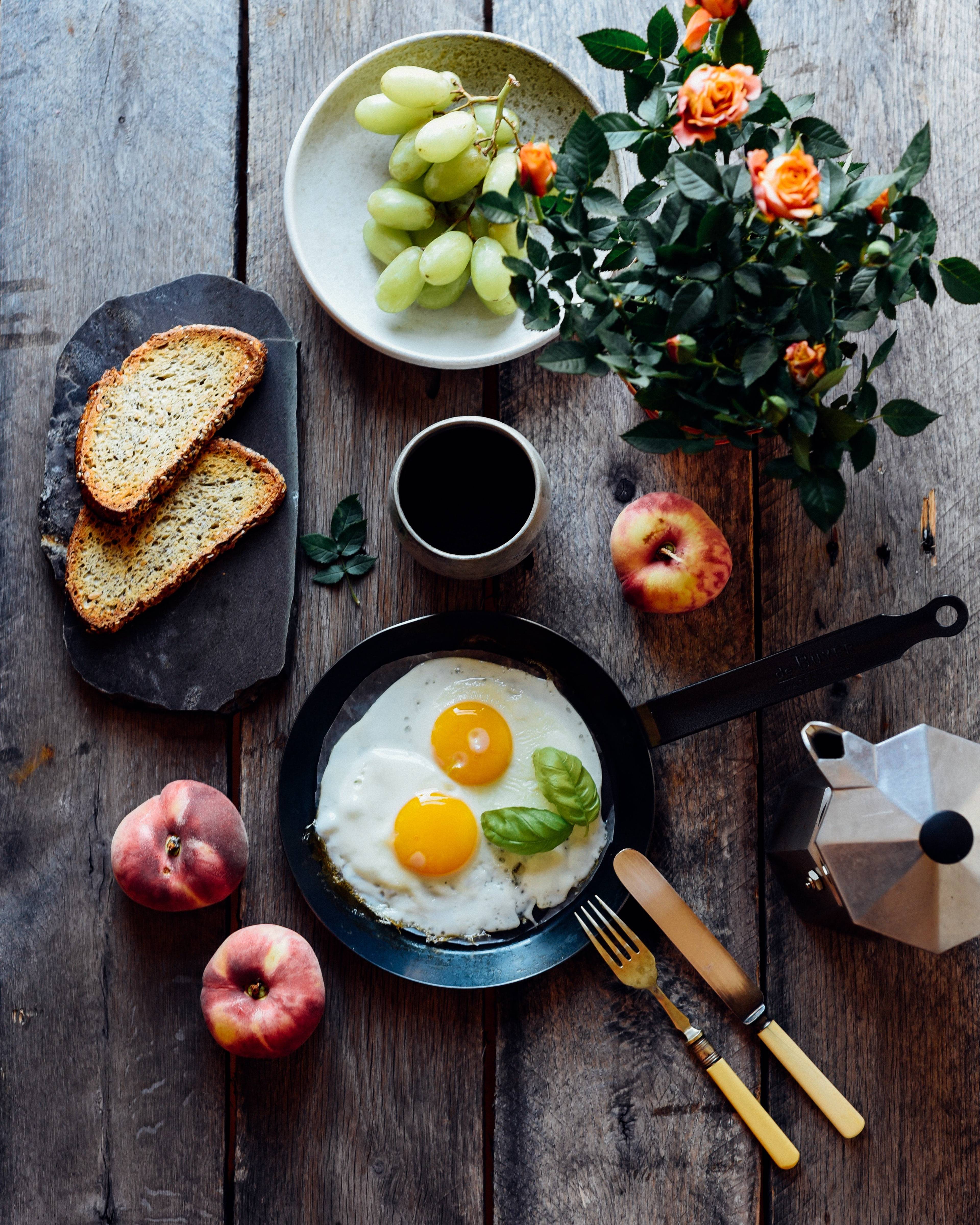 Overhead table with fried eggs, toast, green grapes, peaches, coffee, orange roses, and a moka pot.