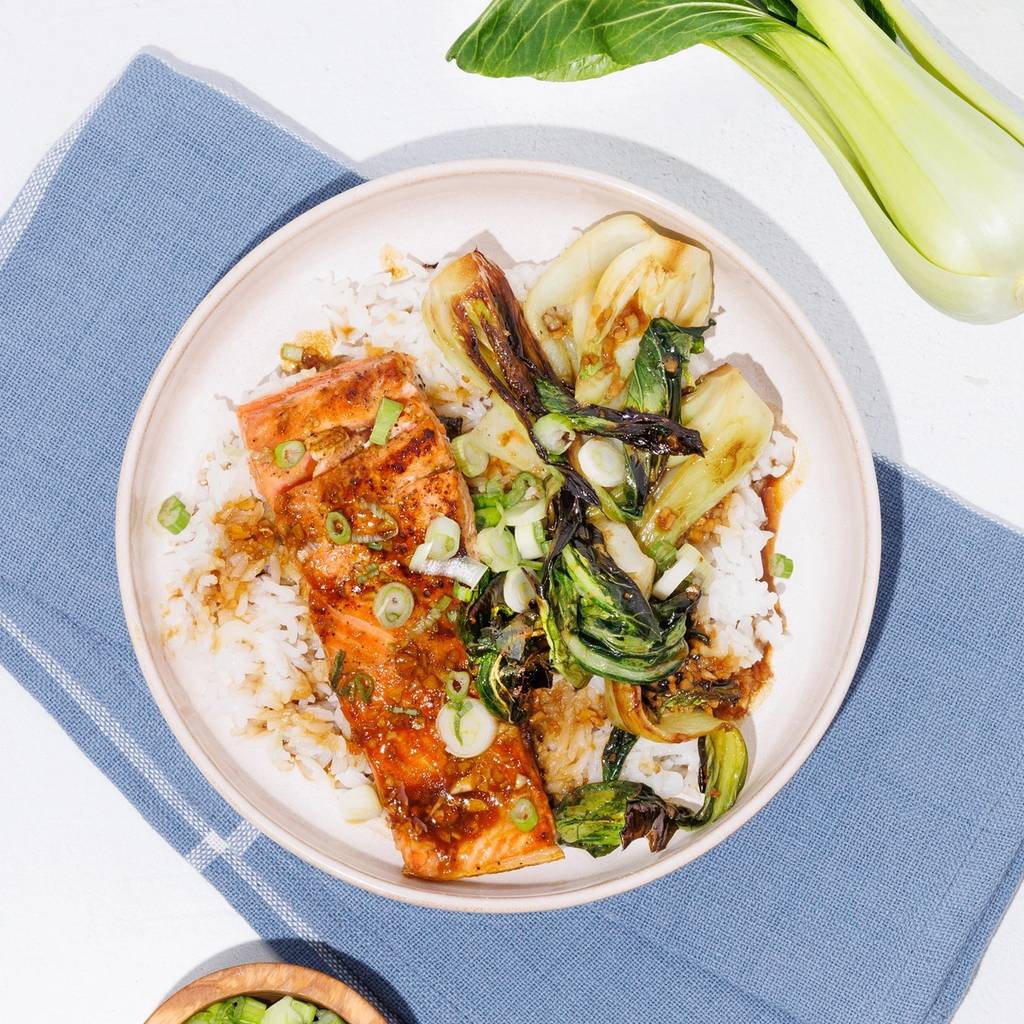 Bowl of glazed salmon, rice, and bok choy with scallions, beside fresh bok choy and garlic.
