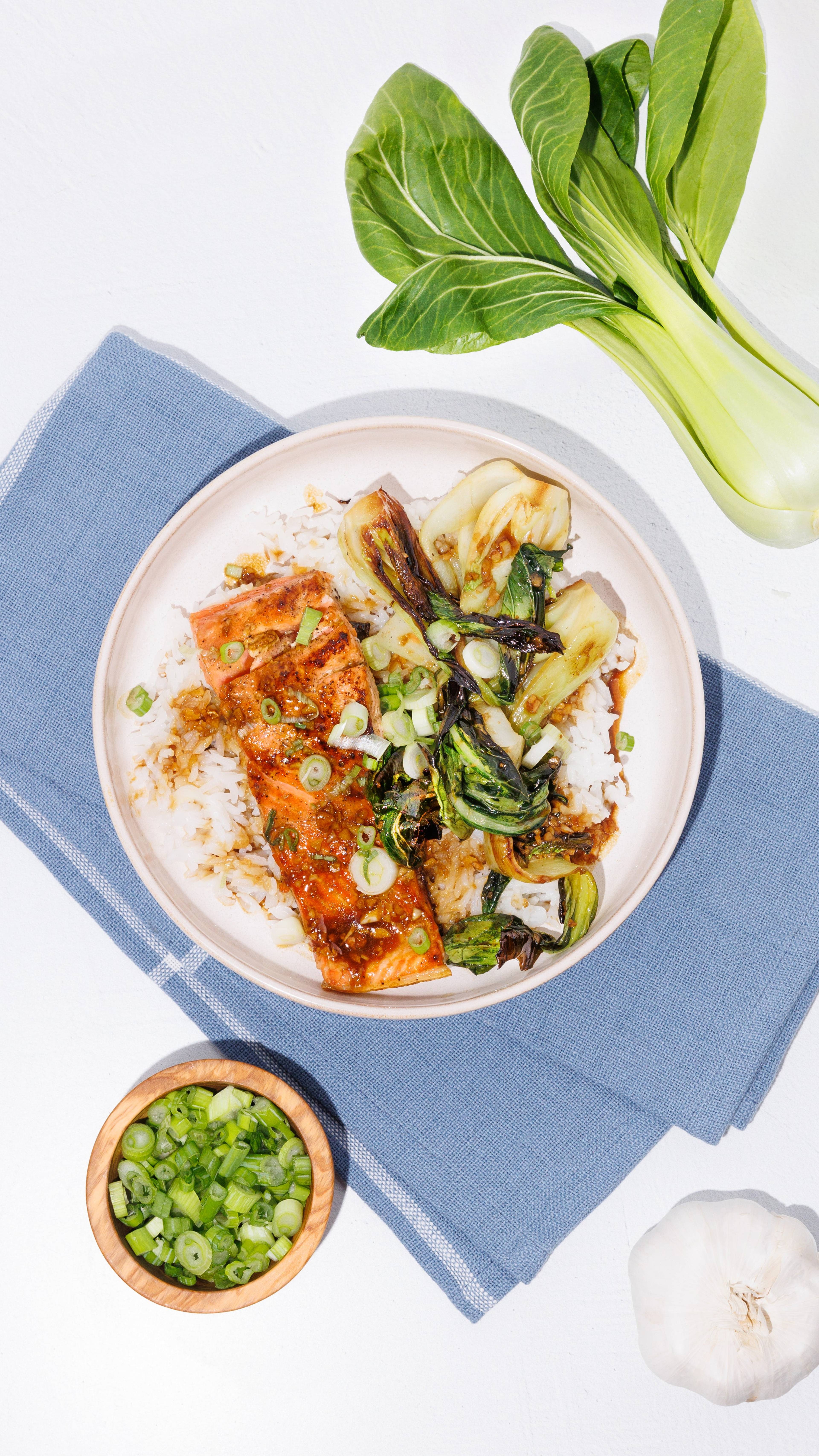 Bowl of glazed salmon, rice, and bok choy with scallions, beside fresh bok choy and garlic.