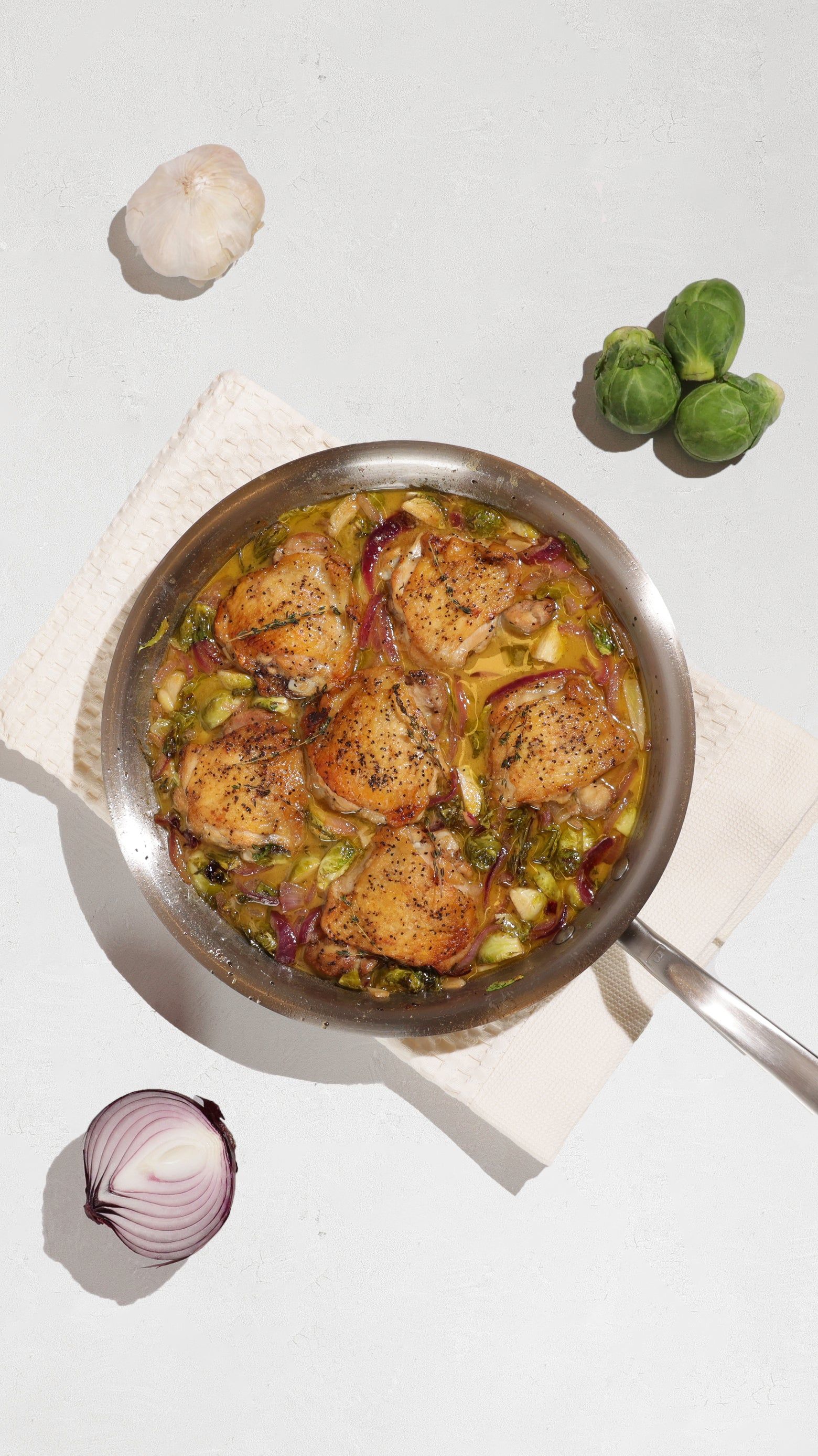 Skillet of browned chicken thighs with Brussels sprouts and red onion, surrounded by garlic and vegetables.