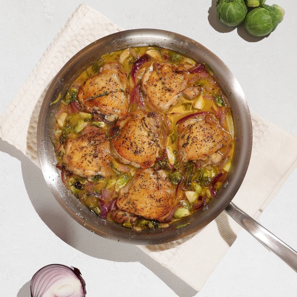 Skillet of browned chicken thighs with Brussels sprouts and red onion, surrounded by garlic and vegetables.