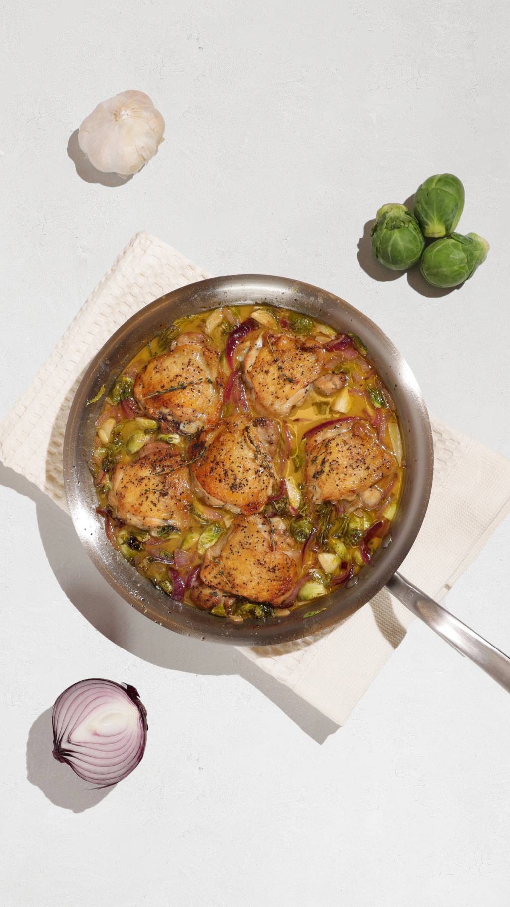 Skillet of browned chicken thighs with Brussels sprouts and red onion, surrounded by garlic and vegetables.