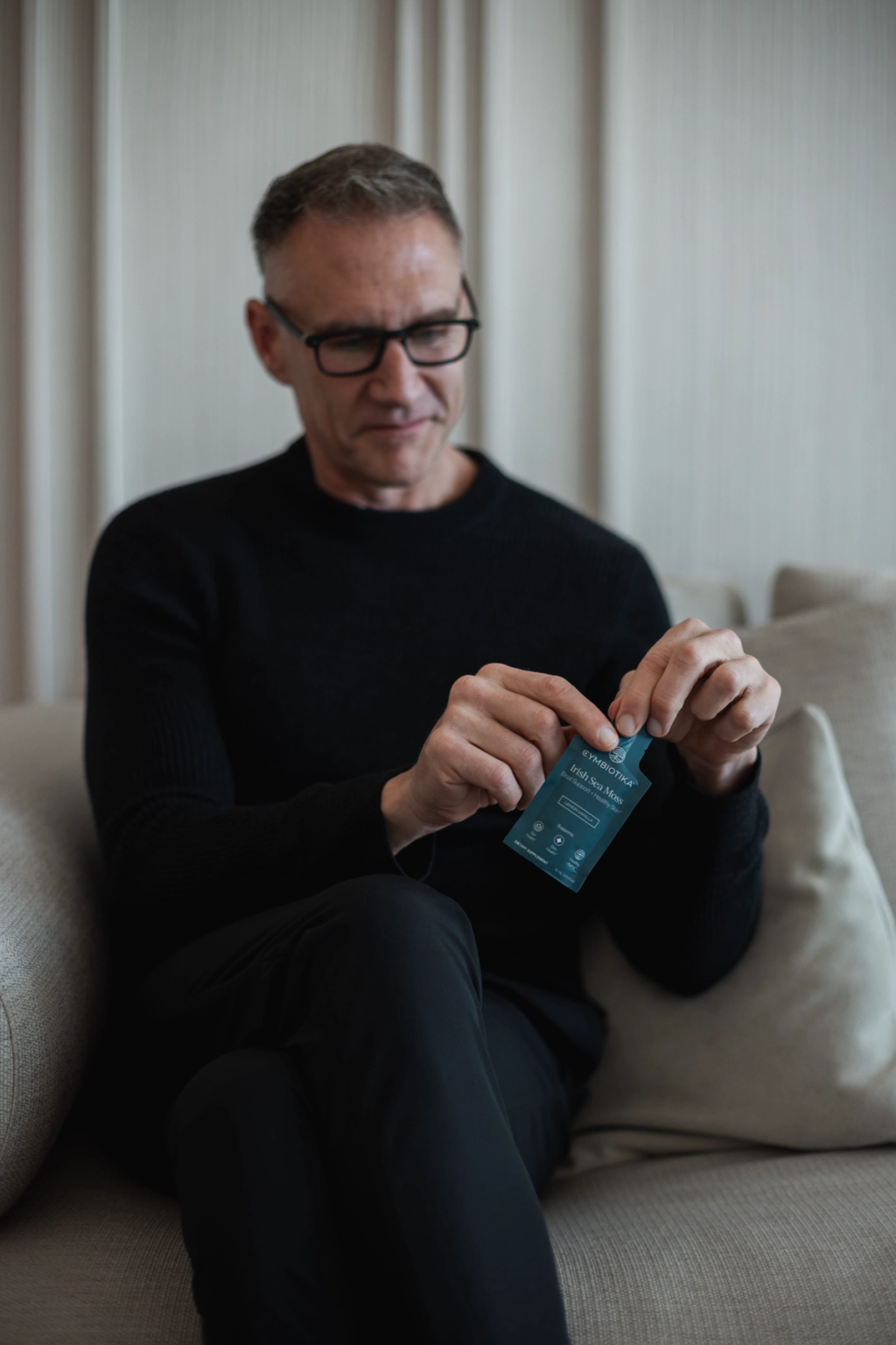 Man in black sweater opening a Cymbiotika Irish Sea Moss sachet while seated on a sofa.