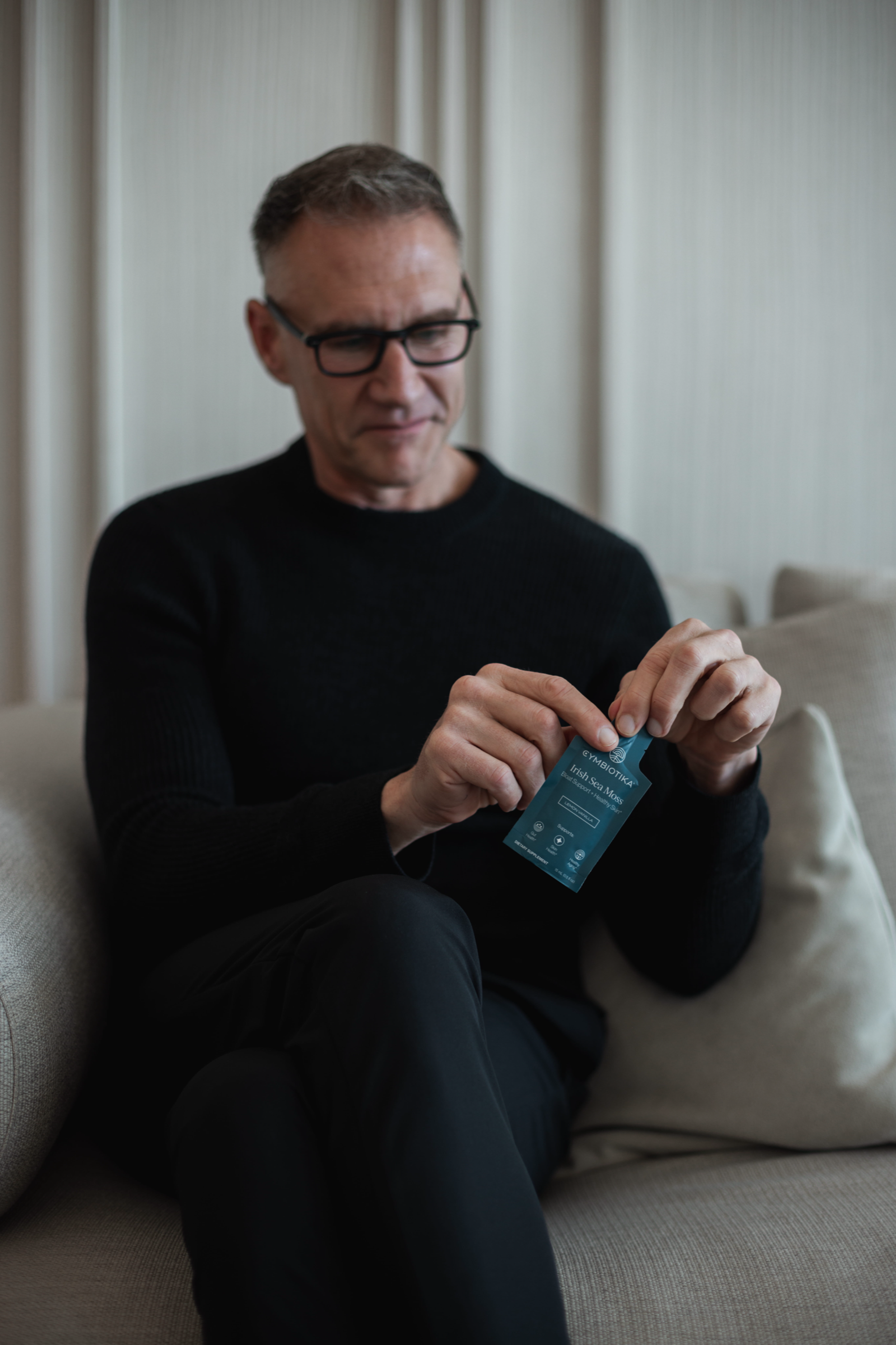 Man in black sweater opening a Cymbiotika Irish Sea Moss sachet while seated on a sofa.