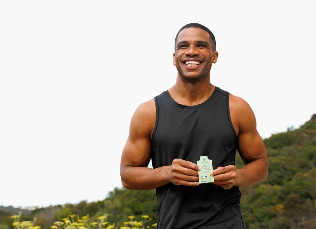 Smiling man in a black tank top holds a small green supplement packet outdoors.