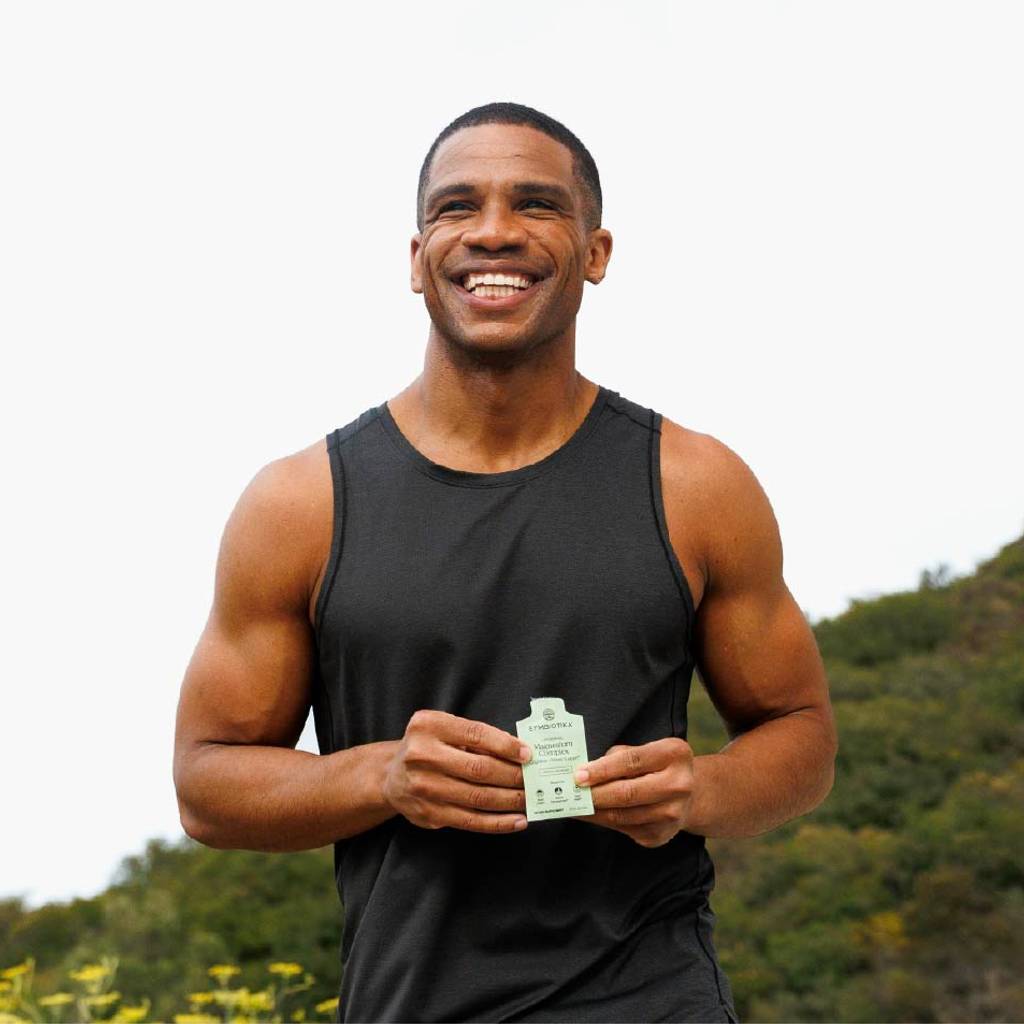 Smiling man in a black tank top holds a small green supplement packet outdoors.