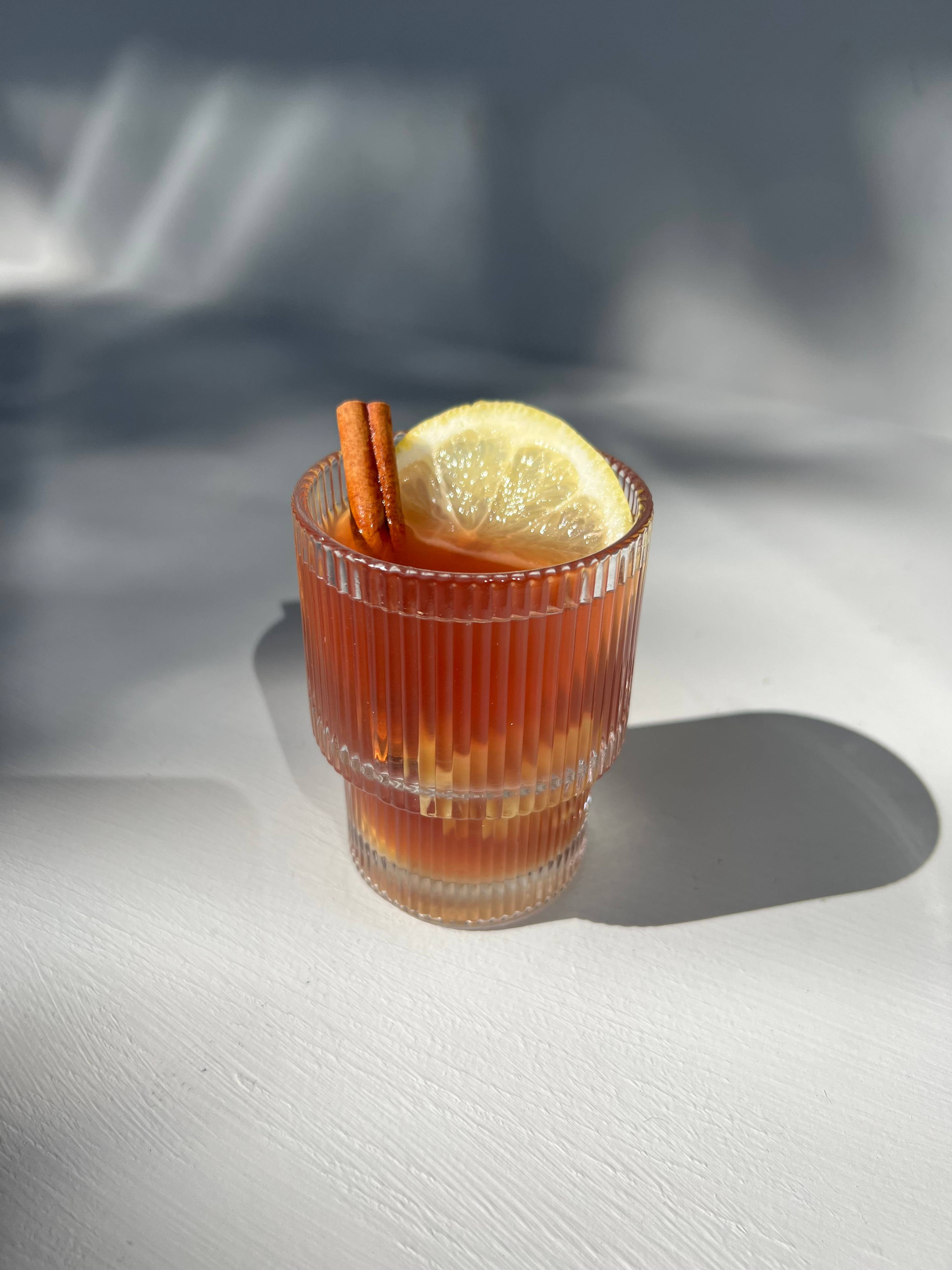 Amber drink garnished with a lemon slice and cinnamon sticks, served in a ribbed glass.