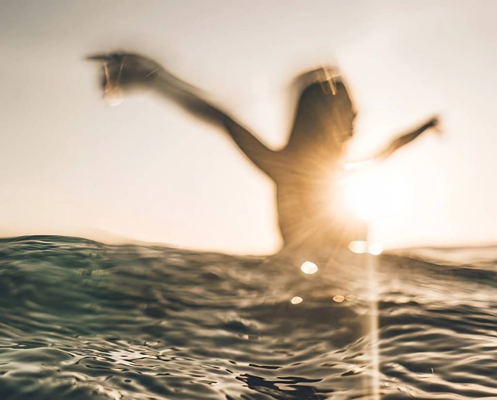 Blurred person with arms raised standing in ocean water at sunset.