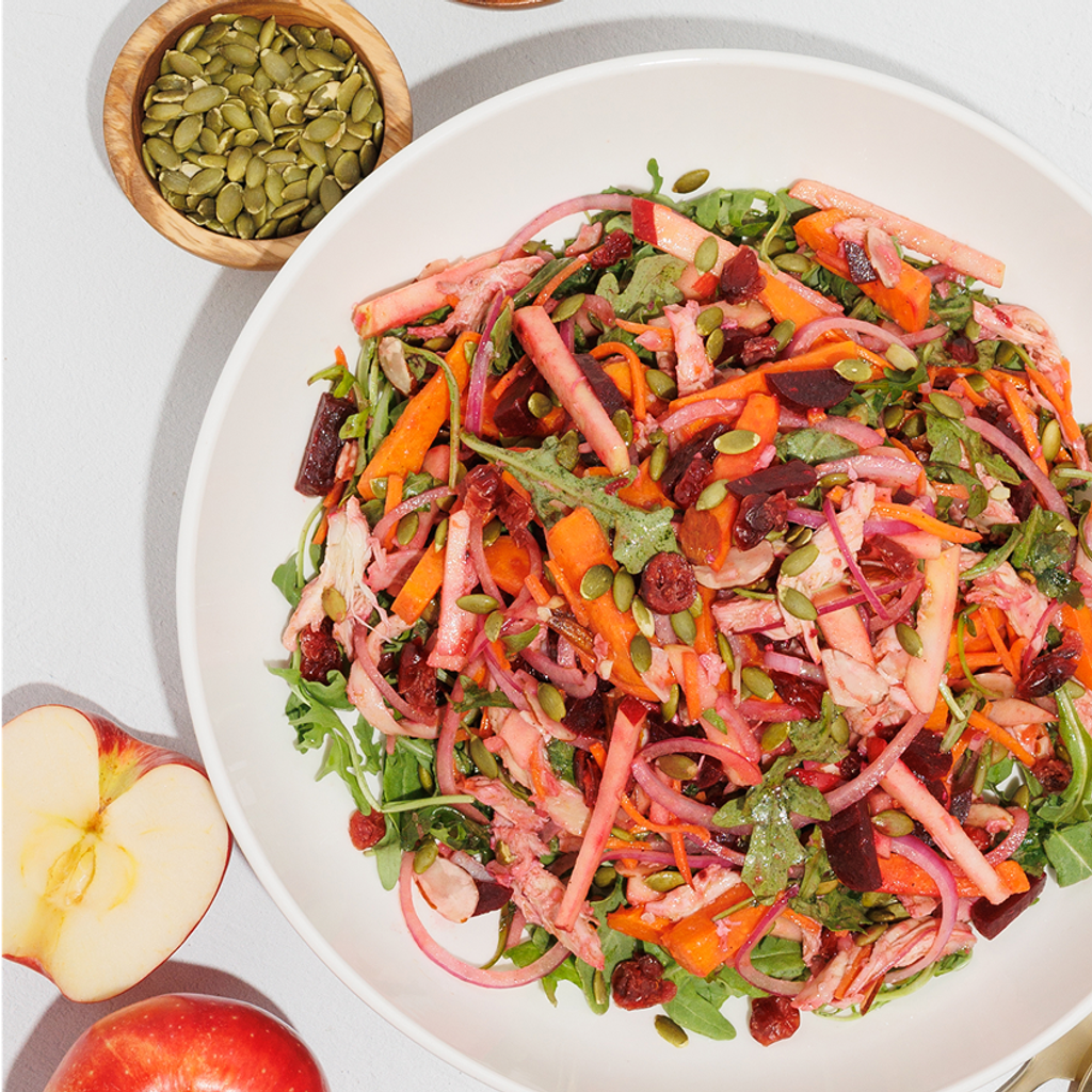 Overhead view of a colorful salad with leafy greens and shredded vegetables, beside sliced apple and a bowl of seeds.