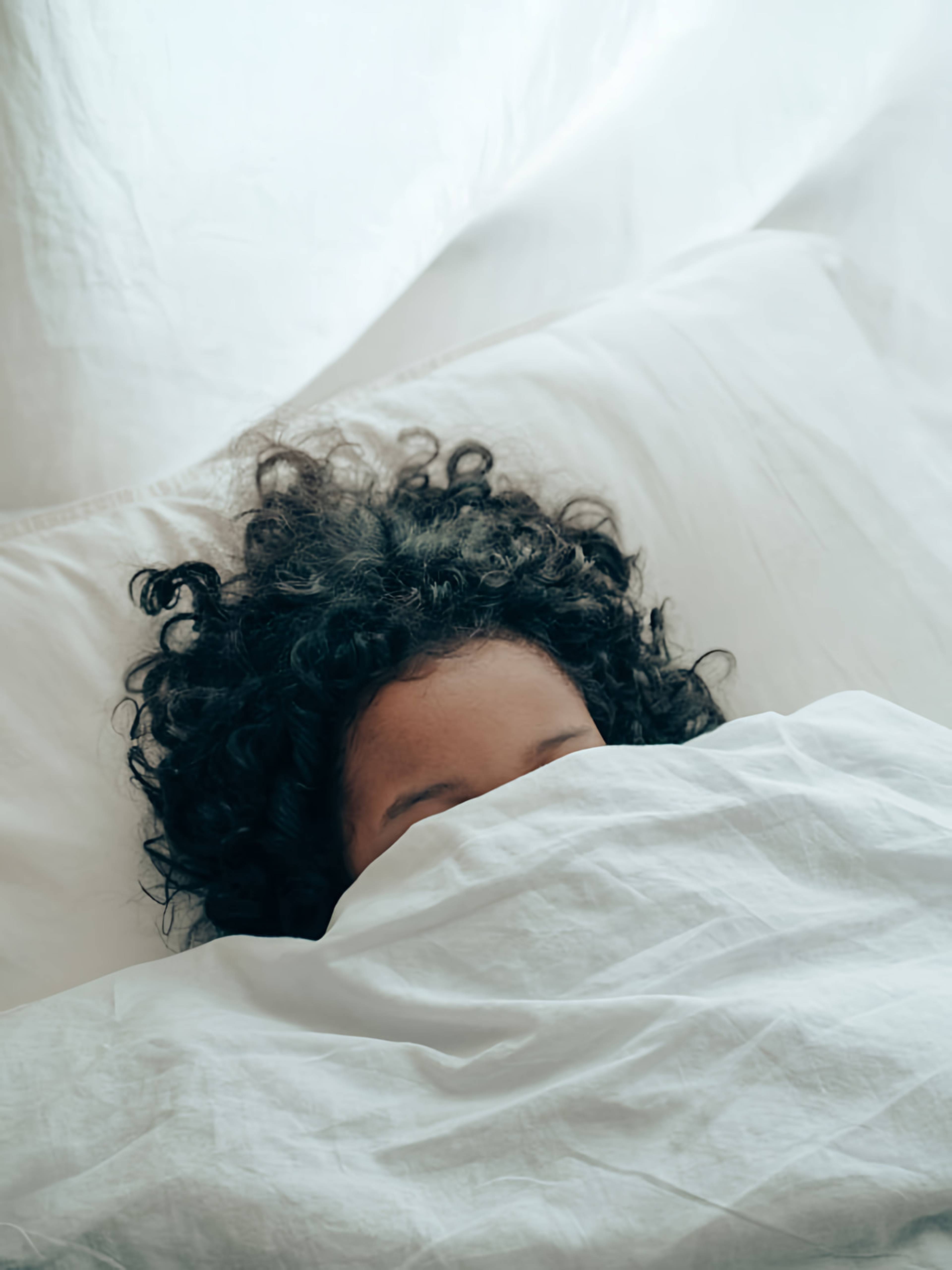 Person with curly dark hair sleeping under a white duvet in bed.