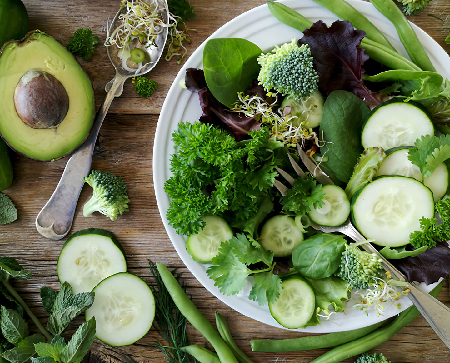Overhead view of a green salad with cucumber, broccoli, leafy greens, sprouts, and avocado on a wooden table.