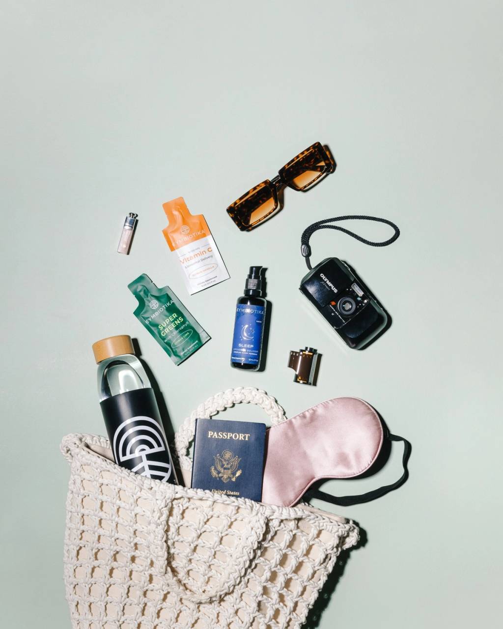 Travel flat lay with a woven bag, water bottle, passport, sleep mask, camera, sunglasses, and Cymbiotika supplements.