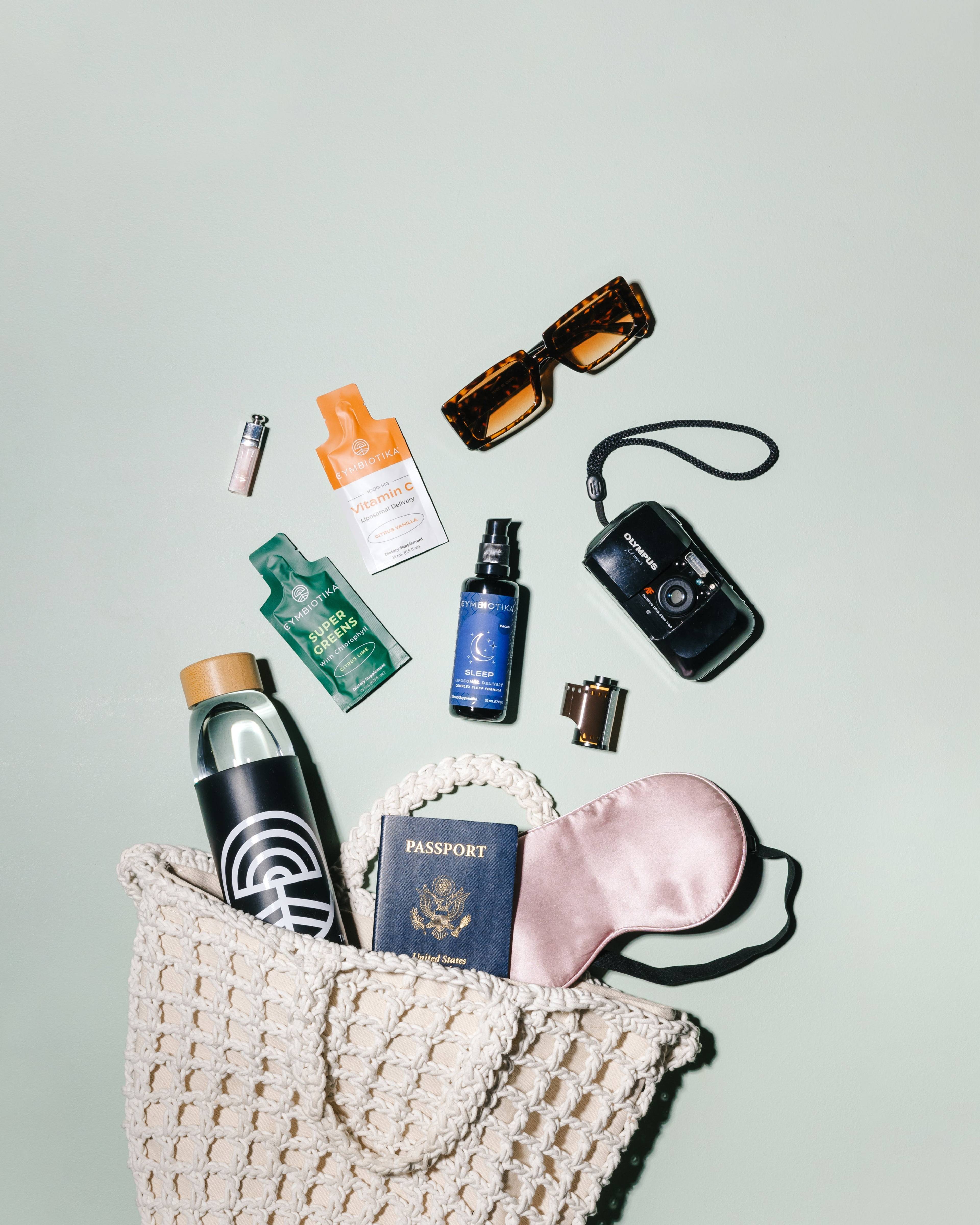 Travel flat lay with a woven bag, water bottle, passport, sleep mask, camera, sunglasses, and Cymbiotika supplements.