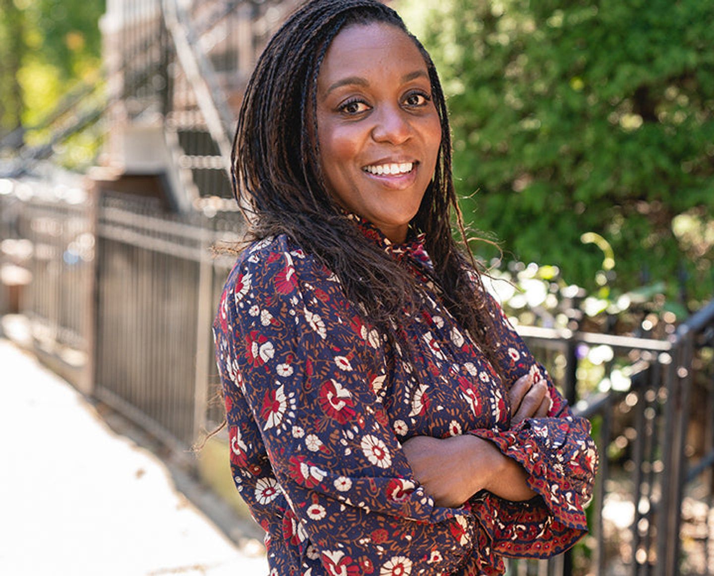 Smiling woman in a floral blouse standing outdoors with her arms crossed.