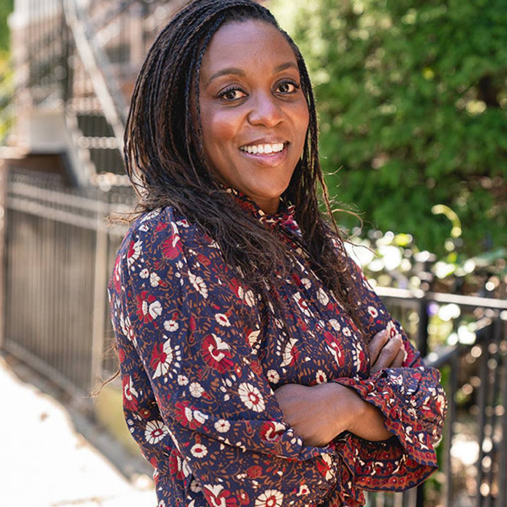 Smiling woman in a floral blouse standing outdoors with her arms crossed.