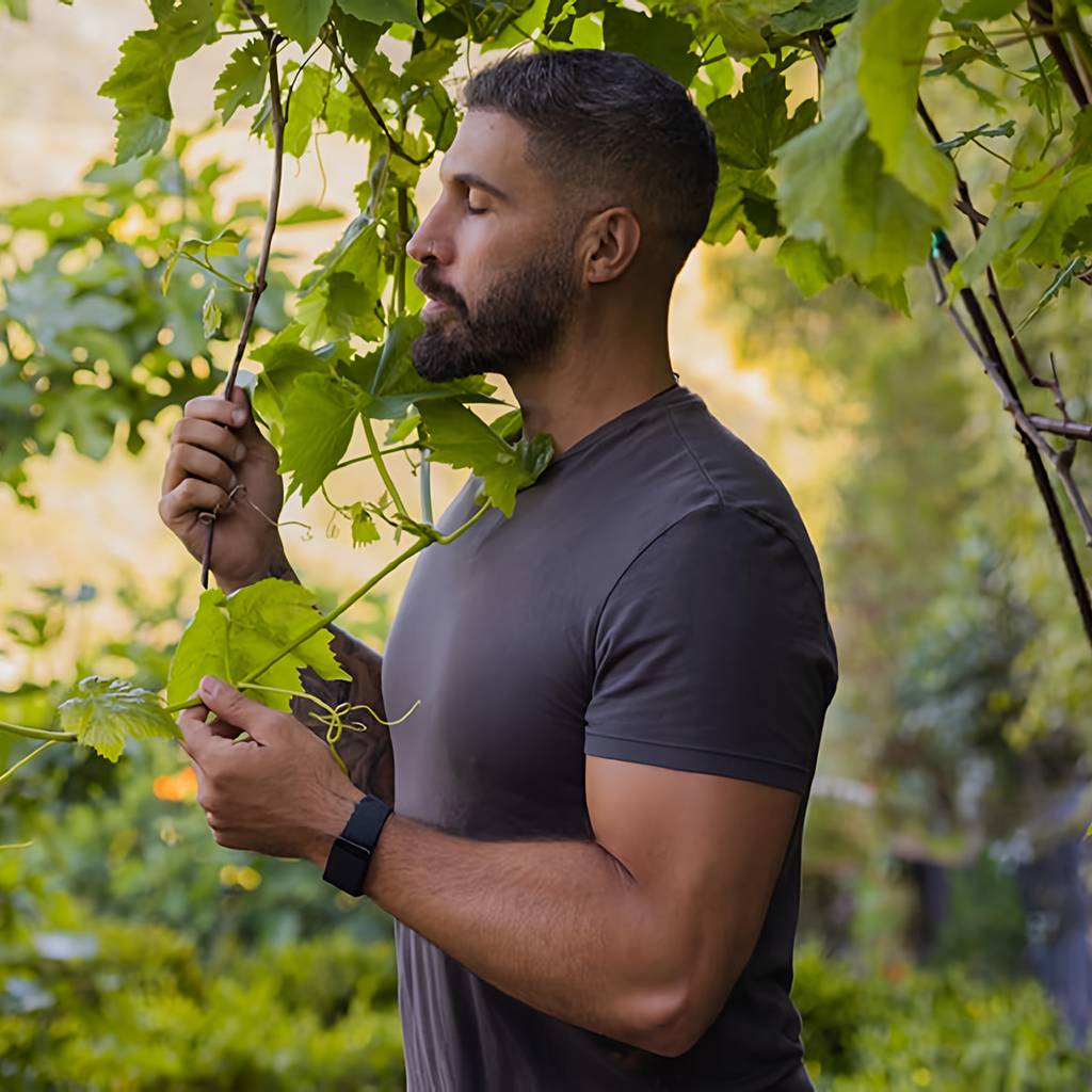 Bearded man in a dark T-shirt standing among leafy vines outdoors.
