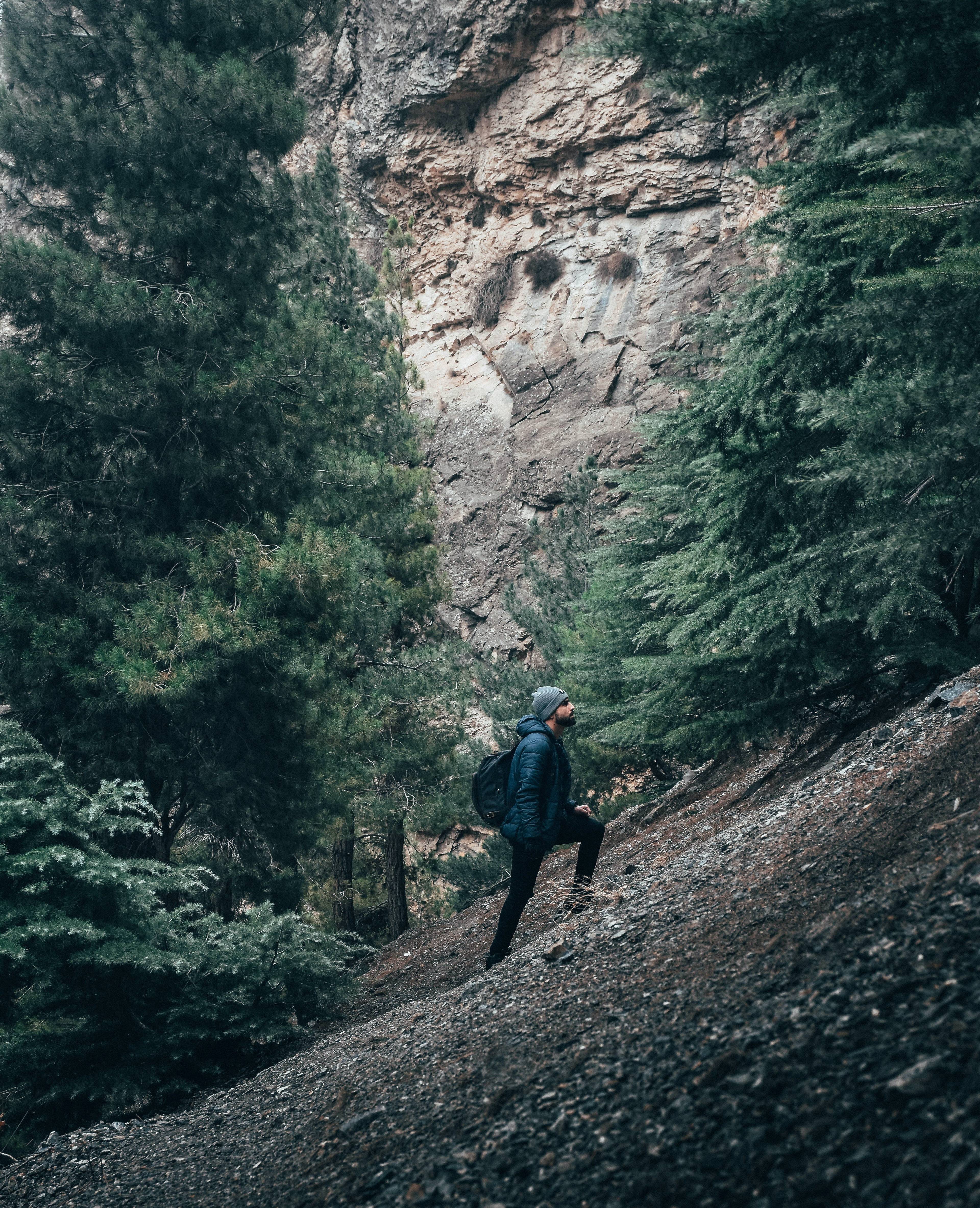 Hiker with a backpack climbing a rocky forest slope below a tall cliff.