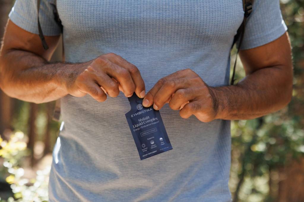Person in a gray shirt opening a Cymbiotika Shilajit Liquid Complex packet outdoors.